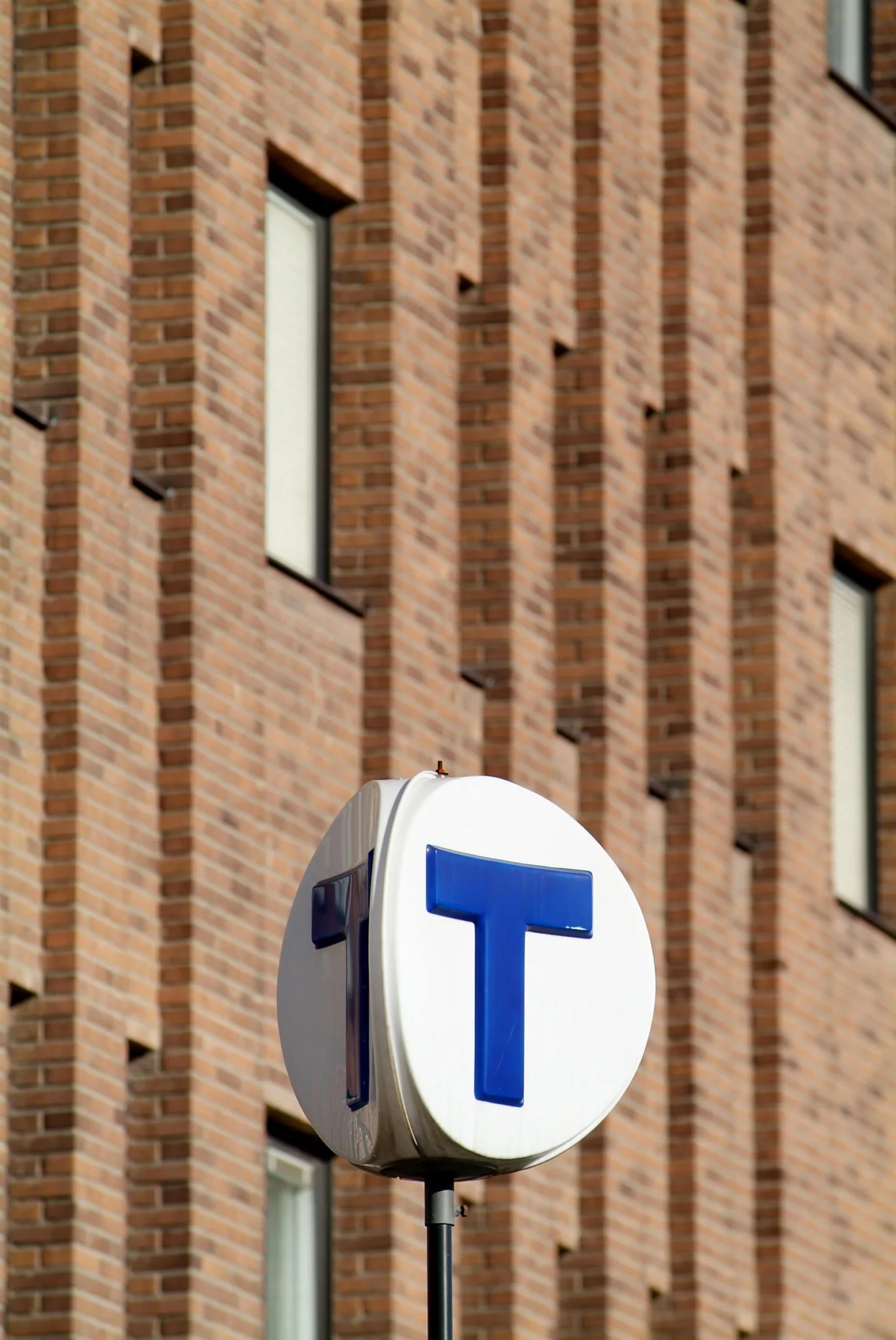En rund skylt med ett blått "T" framför en brun tegelvägg.