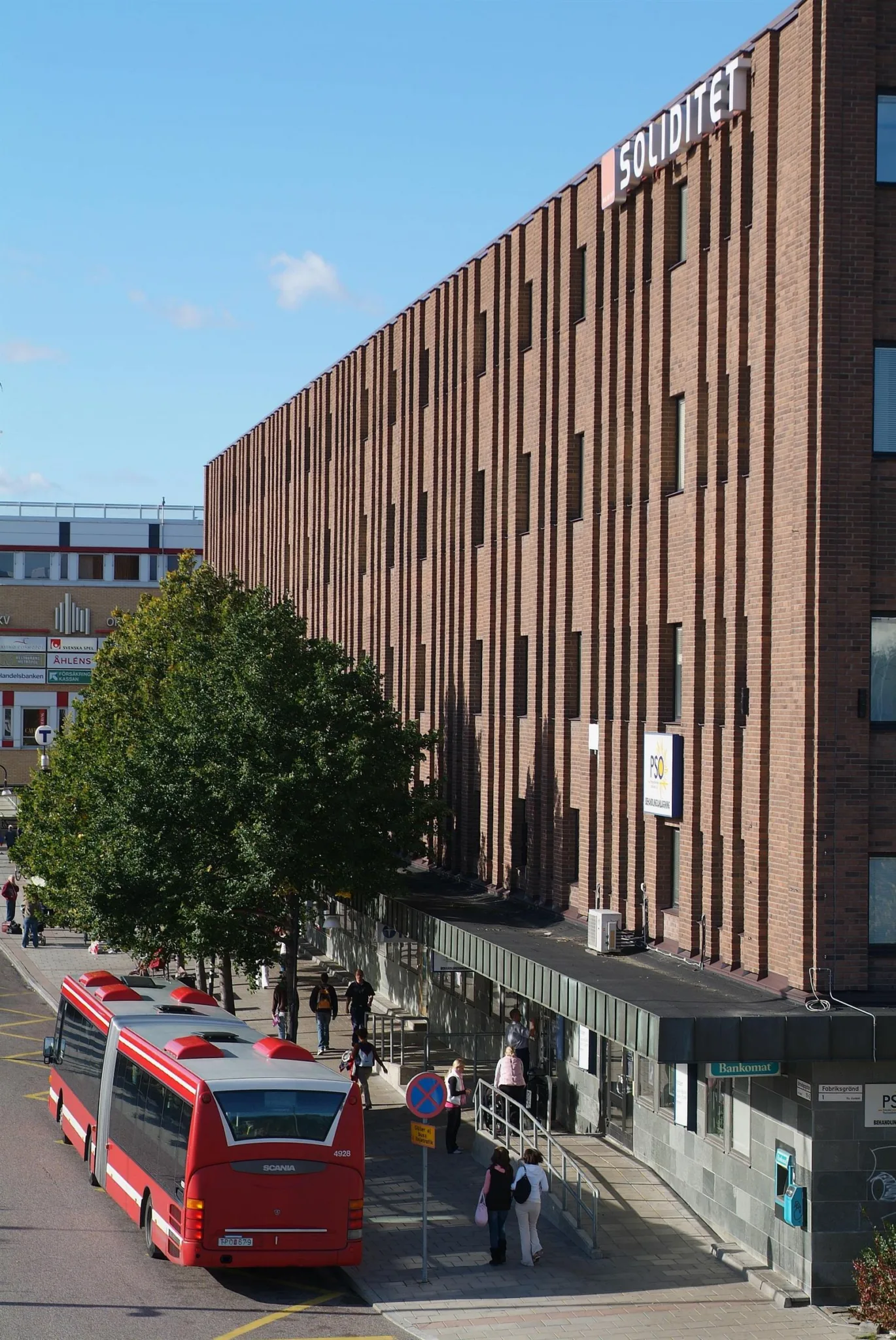 En brun tegelbyggnad med en buss på gatan och människor som promenerar.