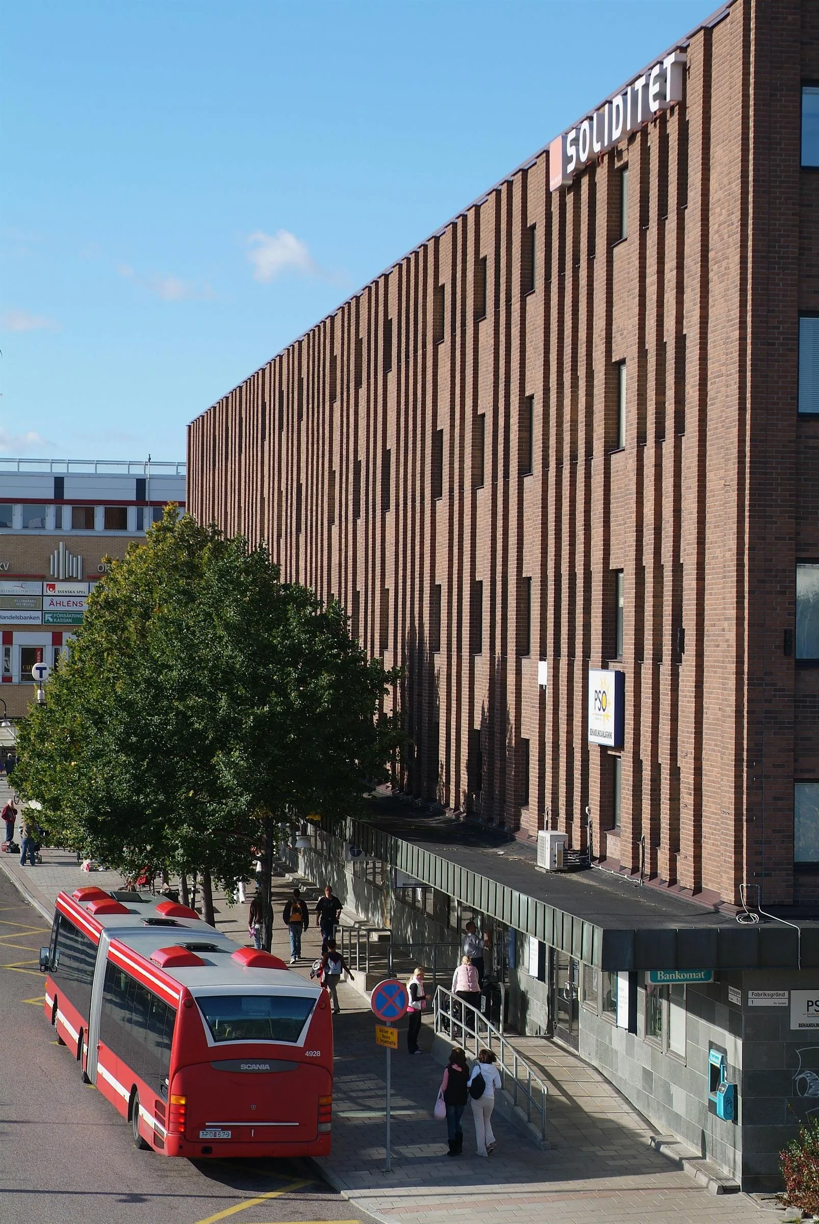 En brun tegelbyggnad med en buss på gatan och människor som promenerar.