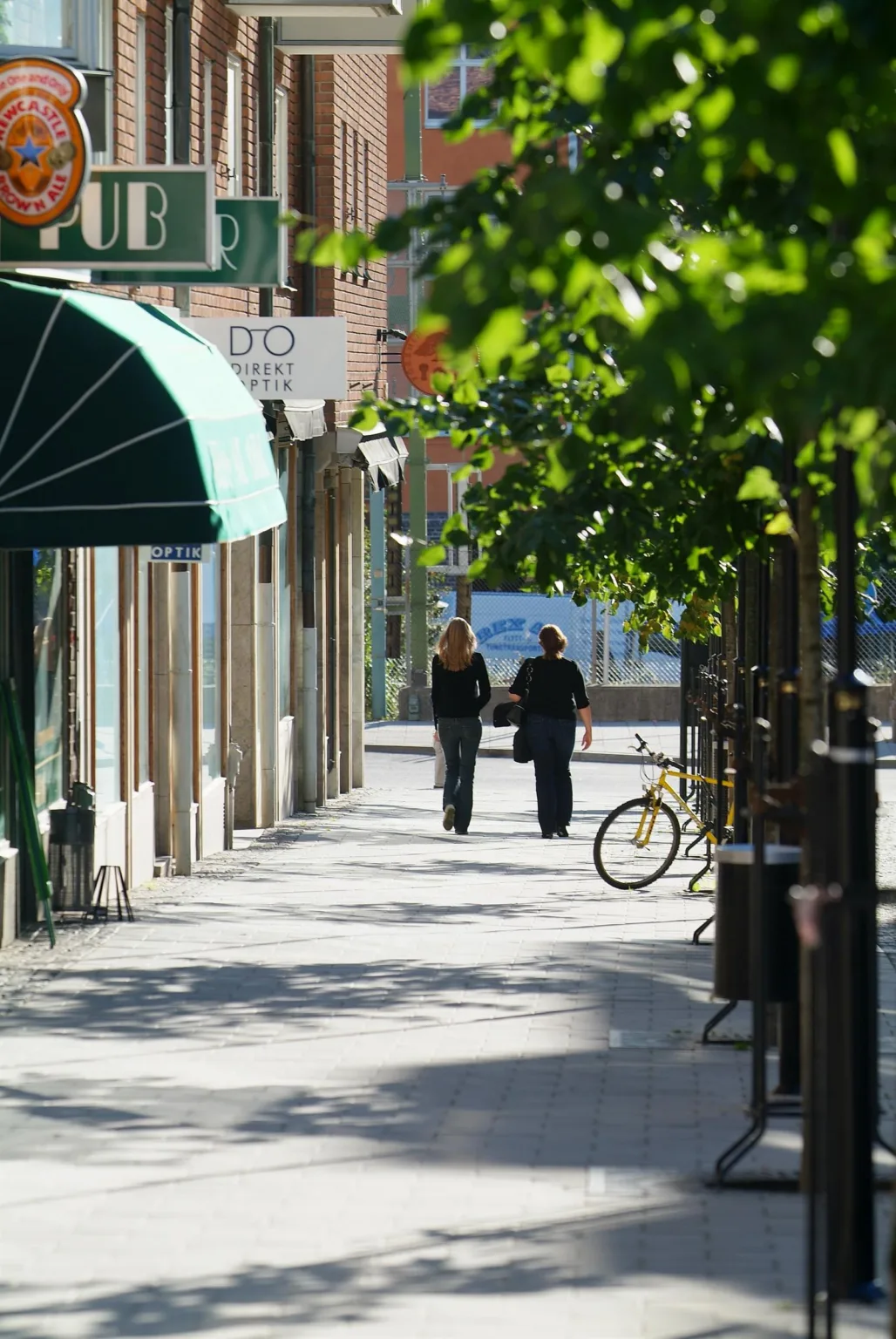 En trottoar kantad av träd med två personer som går, en cykel lutad mot ett staket och affärer i bakgrunden.