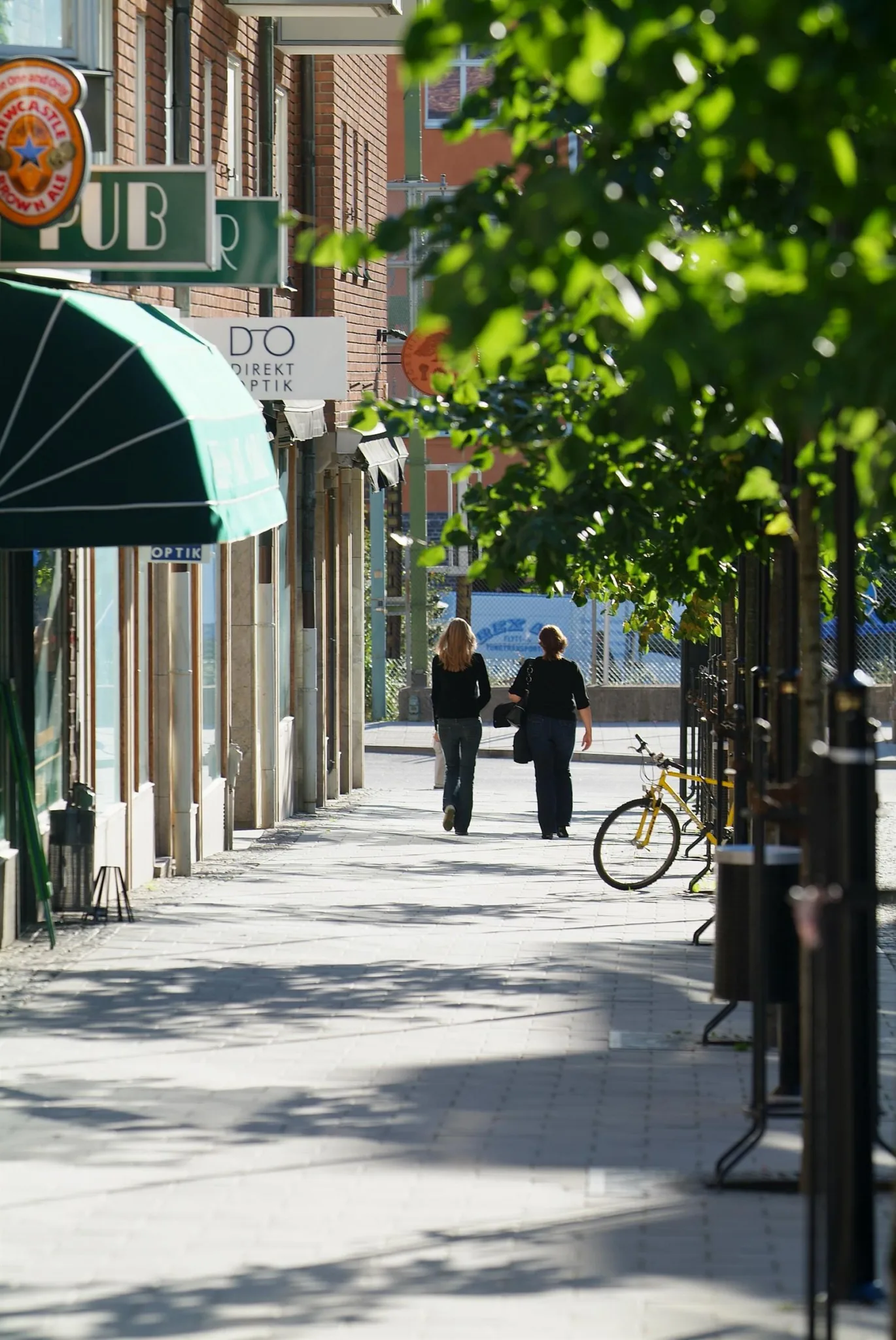 En trottoar kantad av träd med två personer som går, en cykel lutad mot ett staket och affärer i bakgrunden.