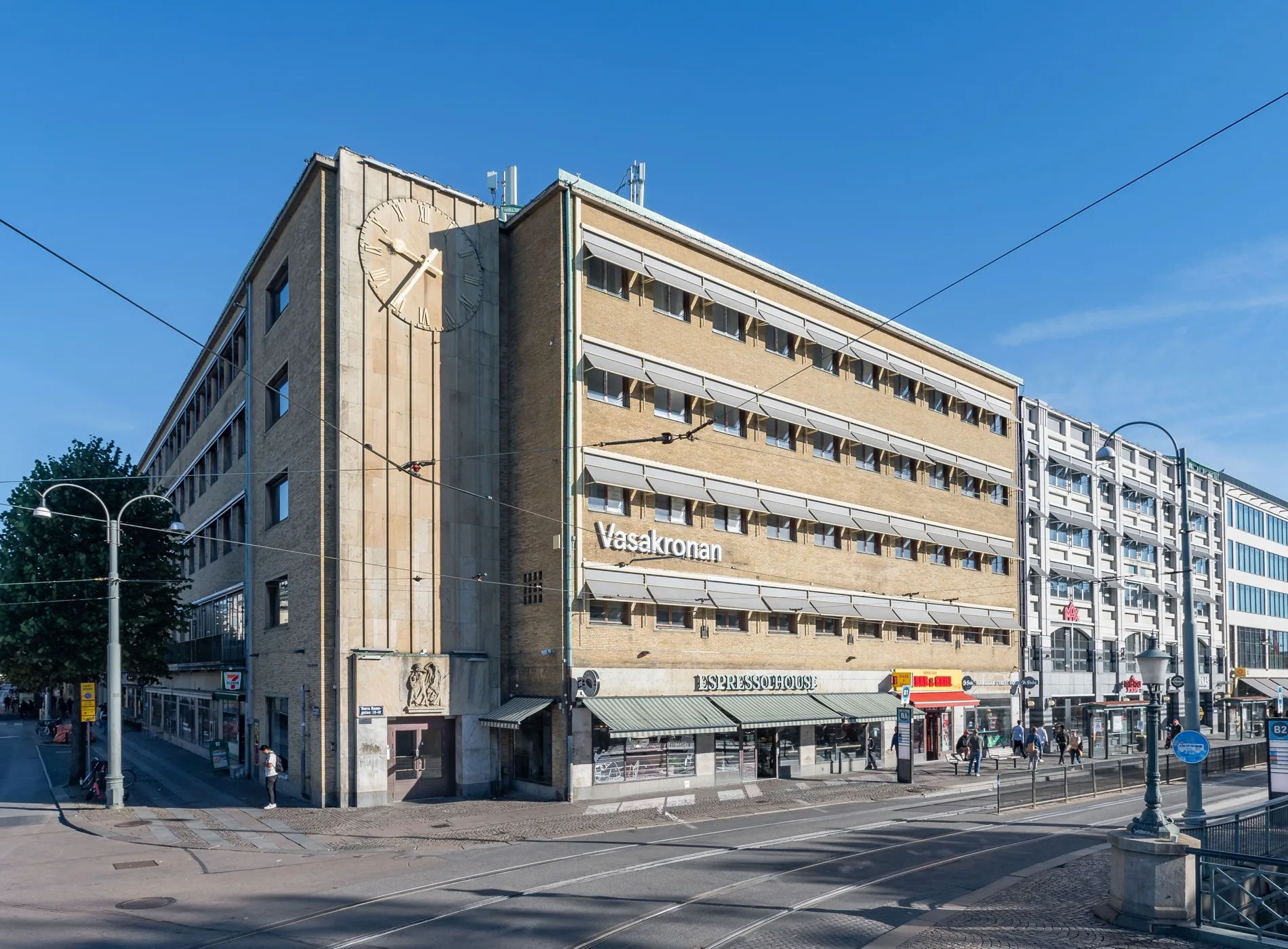 En gul byggnad med flera våningar och butiker vid en korsning under en molnfri himmel.