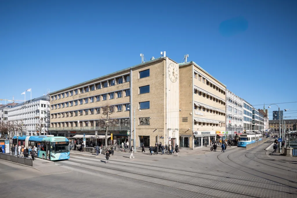 En stor byggnad vid en korsning, omgiven av spårvagnar och fotgängare under en klarblå himmel.
