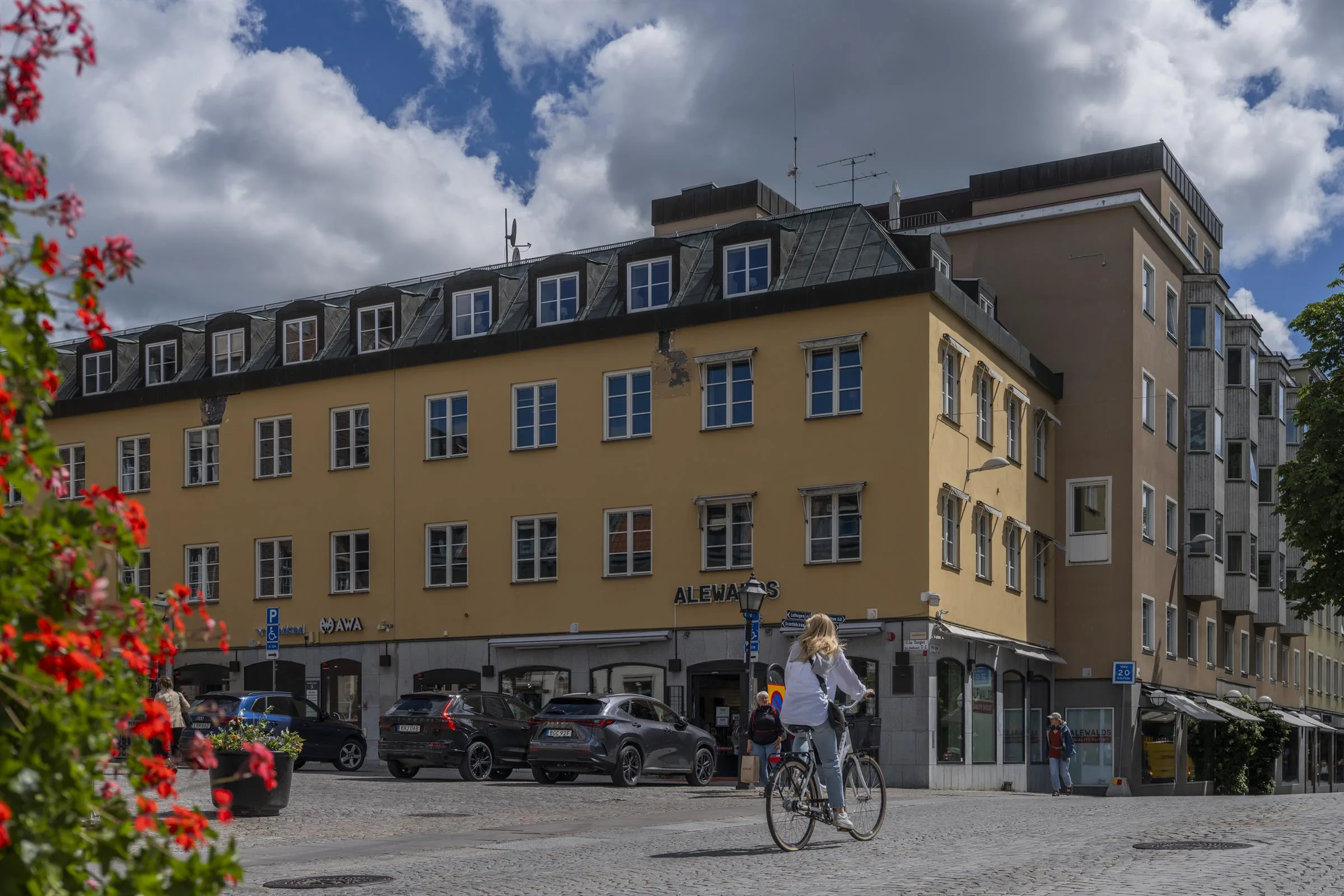 En gul byggnad med parkerade bilar och en kvinna som cyklar, himlen är blå med vita moln.