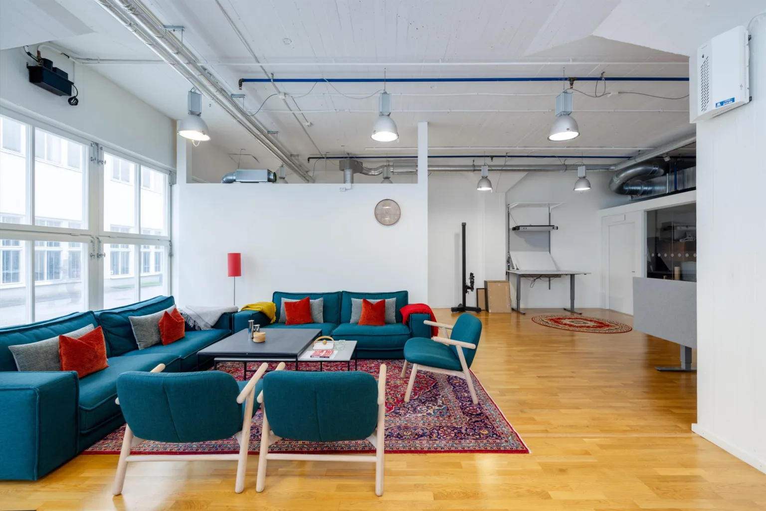 En lounge med blå soffor, röda kuddar och ett färgglatt matta på trägolv, med industriell inredning.