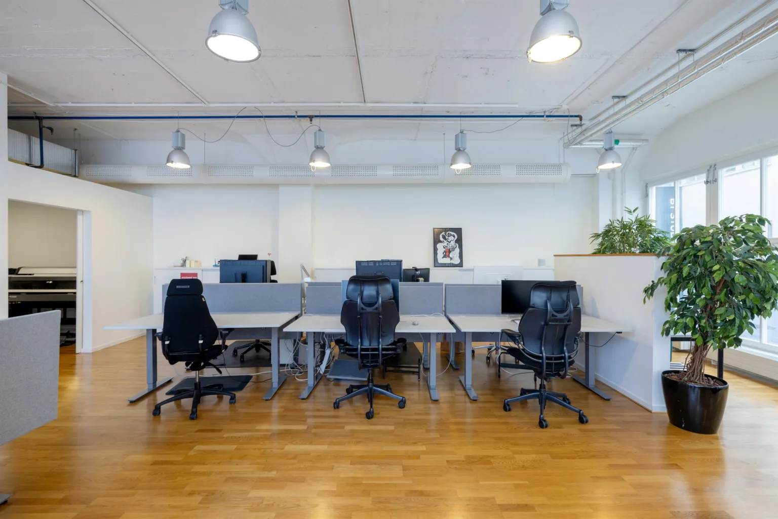 En arbetsplats med flera skrivbord, svarta kontorsstolar och moderna lampor i ett rymligt kontorsutrymme.