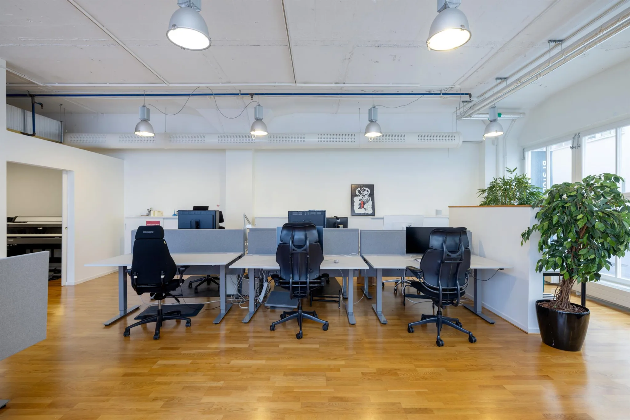 En arbetsplats med flera skrivbord, svarta kontorsstolar och moderna lampor i ett rymligt kontorsutrymme.