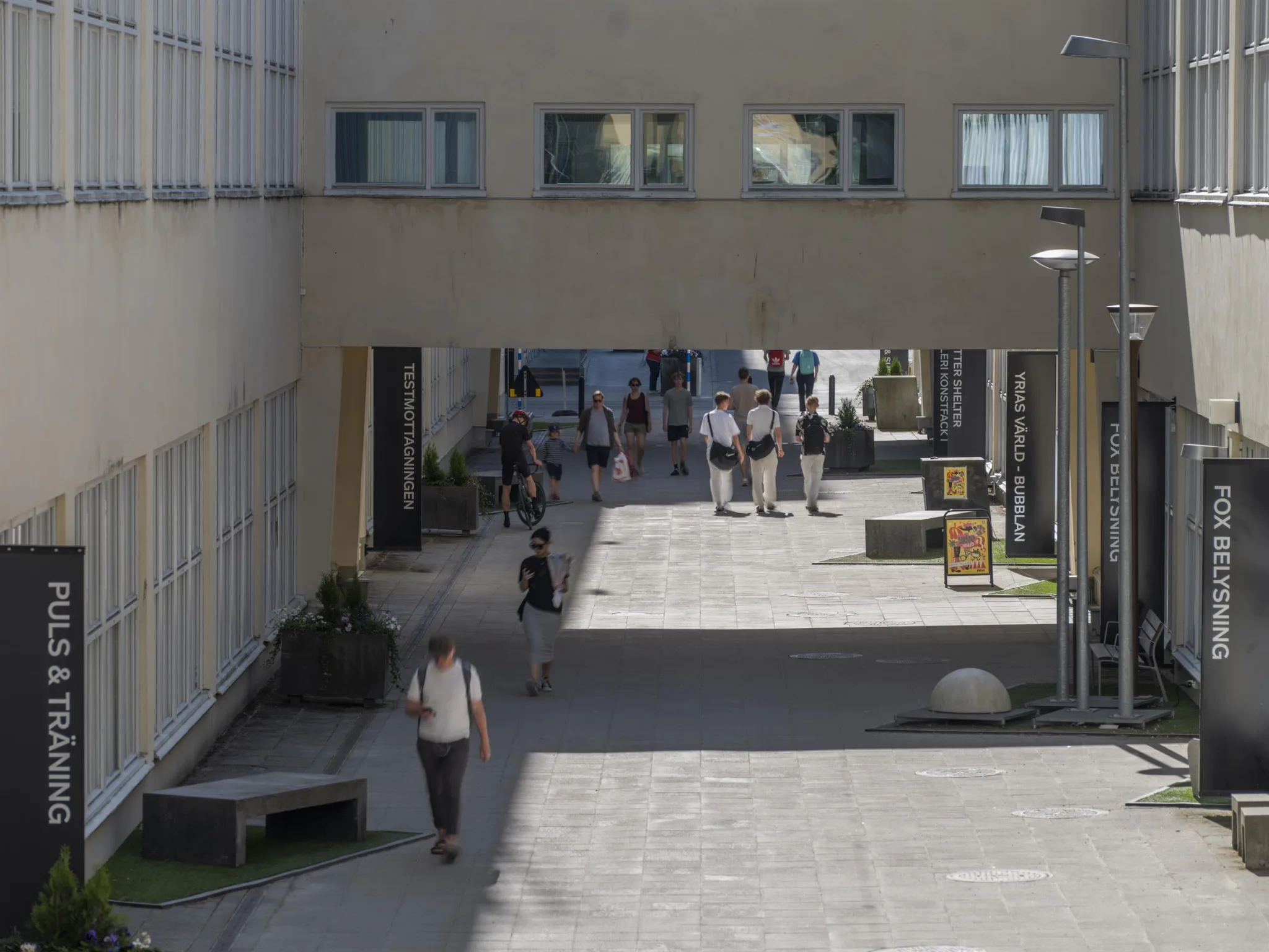 En gångväg mellan byggnader med människor som går och sitter under en öppen passage.