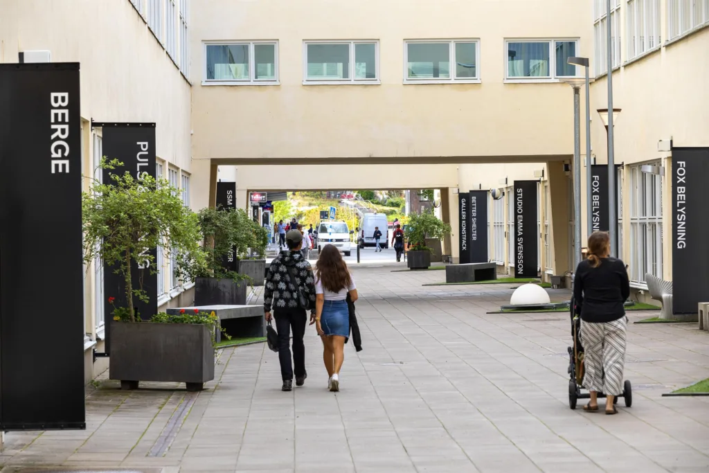 En gångväg mellan byggnader med två personer som går tillsammans och planteringslådor längs sidorna.