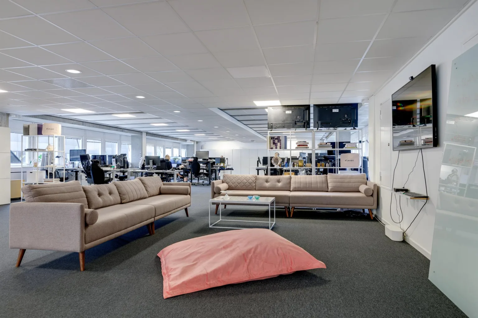Ett öppet kontorsutrymme med soffor, ett rosa sittpuff och arbetsplatser i bakgrunden.