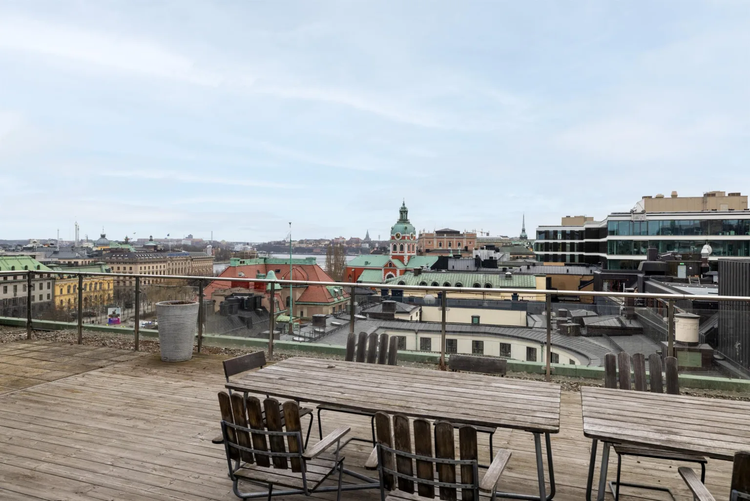 5. En takterrass med träbord och stolar, omgiven av glasräcken och med utsikt över stadens byggnader och kyrktorn.