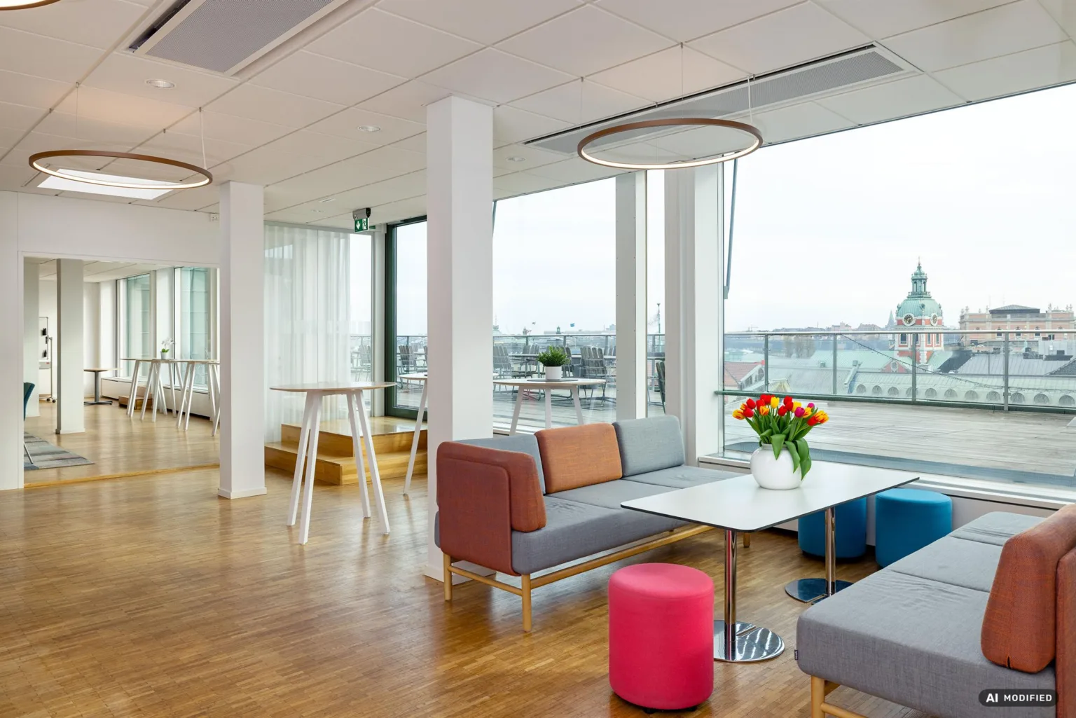 1. En modern lounge med soffor, bord och färgglada sittpuffar, stora fönster med stadsutsikt och en vas med blommor på ett bord.