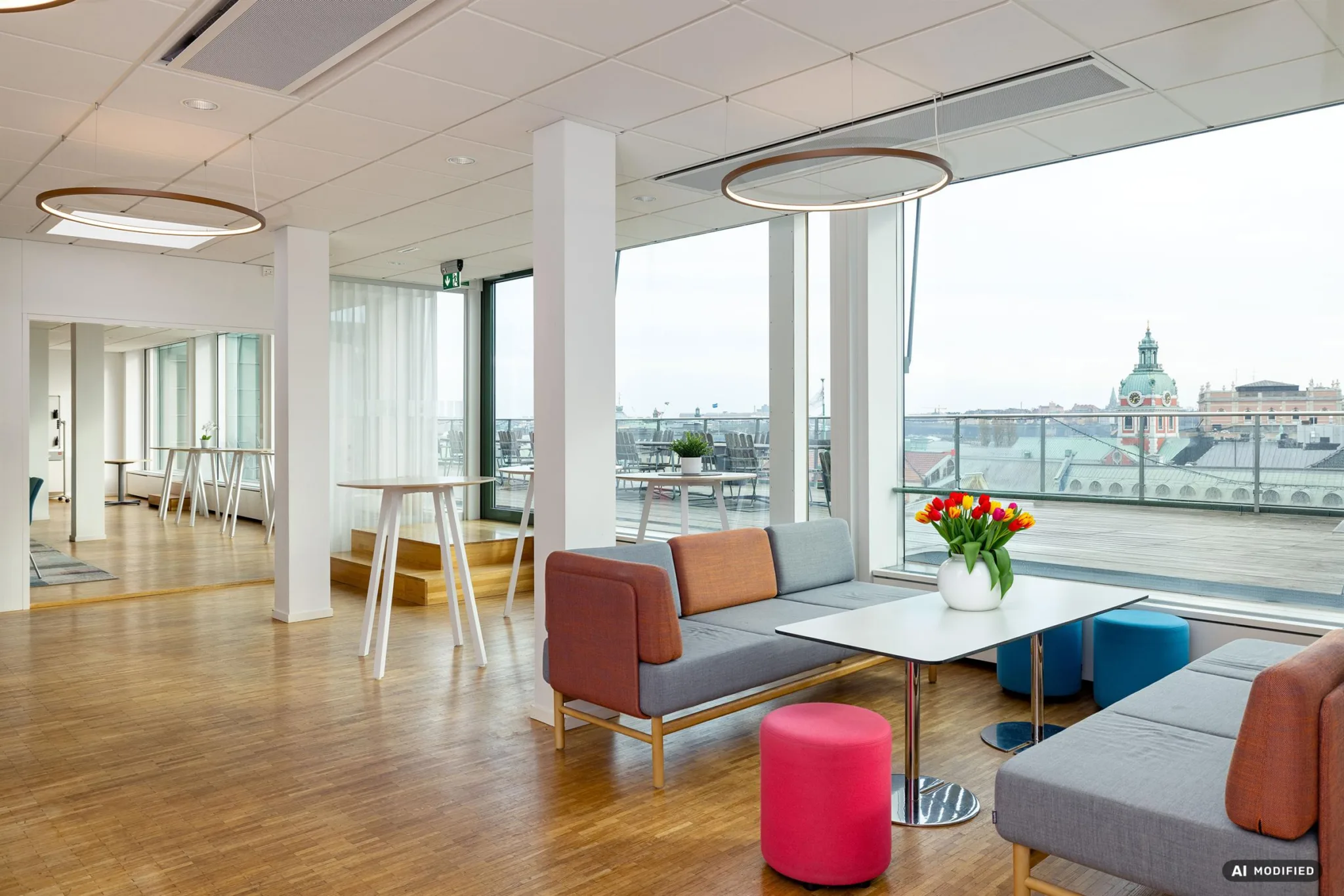 1. En modern lounge med soffor, bord och färgglada sittpuffar, stora fönster med stadsutsikt och en vas med blommor på ett bord.