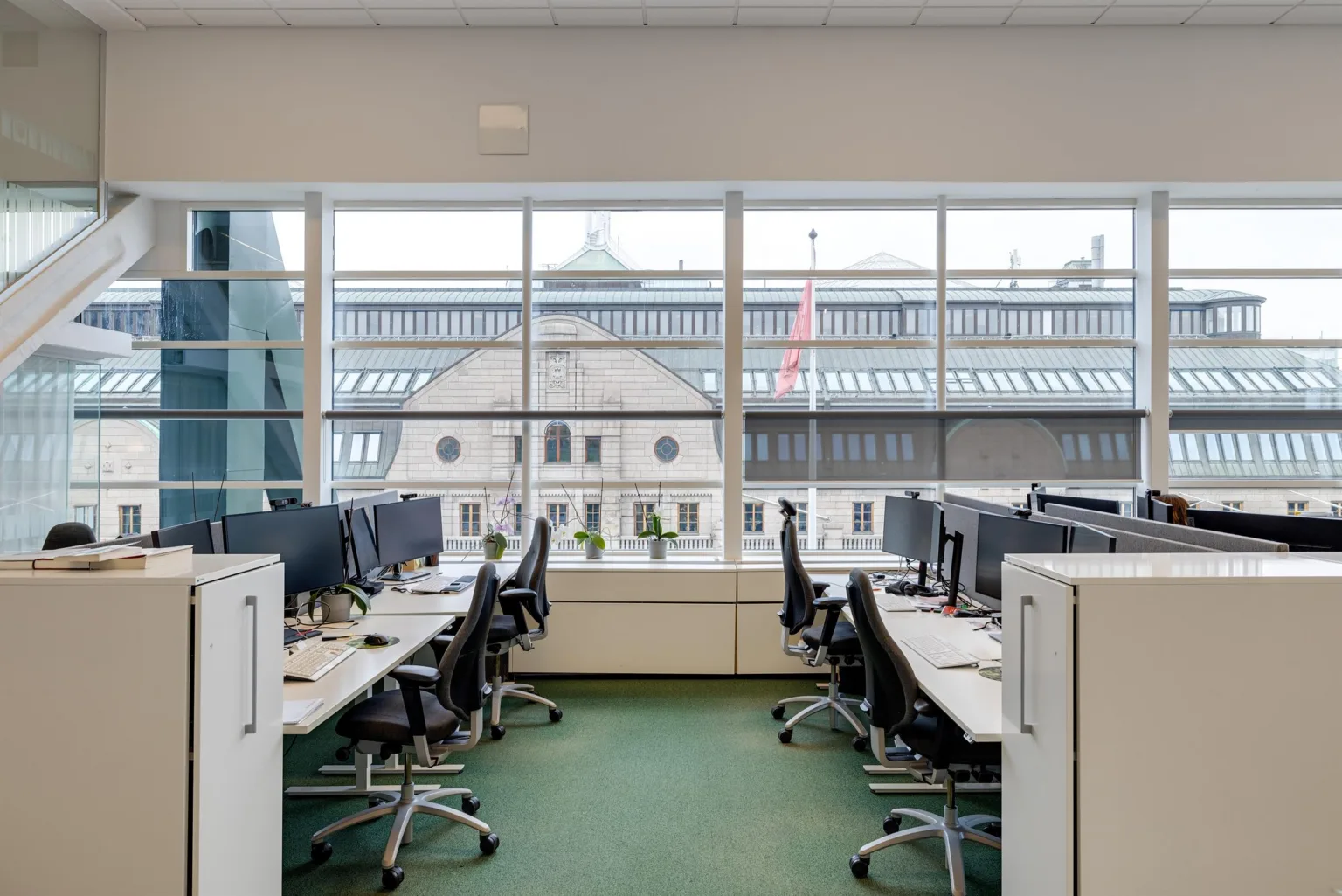 4. Ett kontor med rader av skrivbord och datorer, stora fönster som släpper in dagsljus och utsikt över en byggnad utanför.