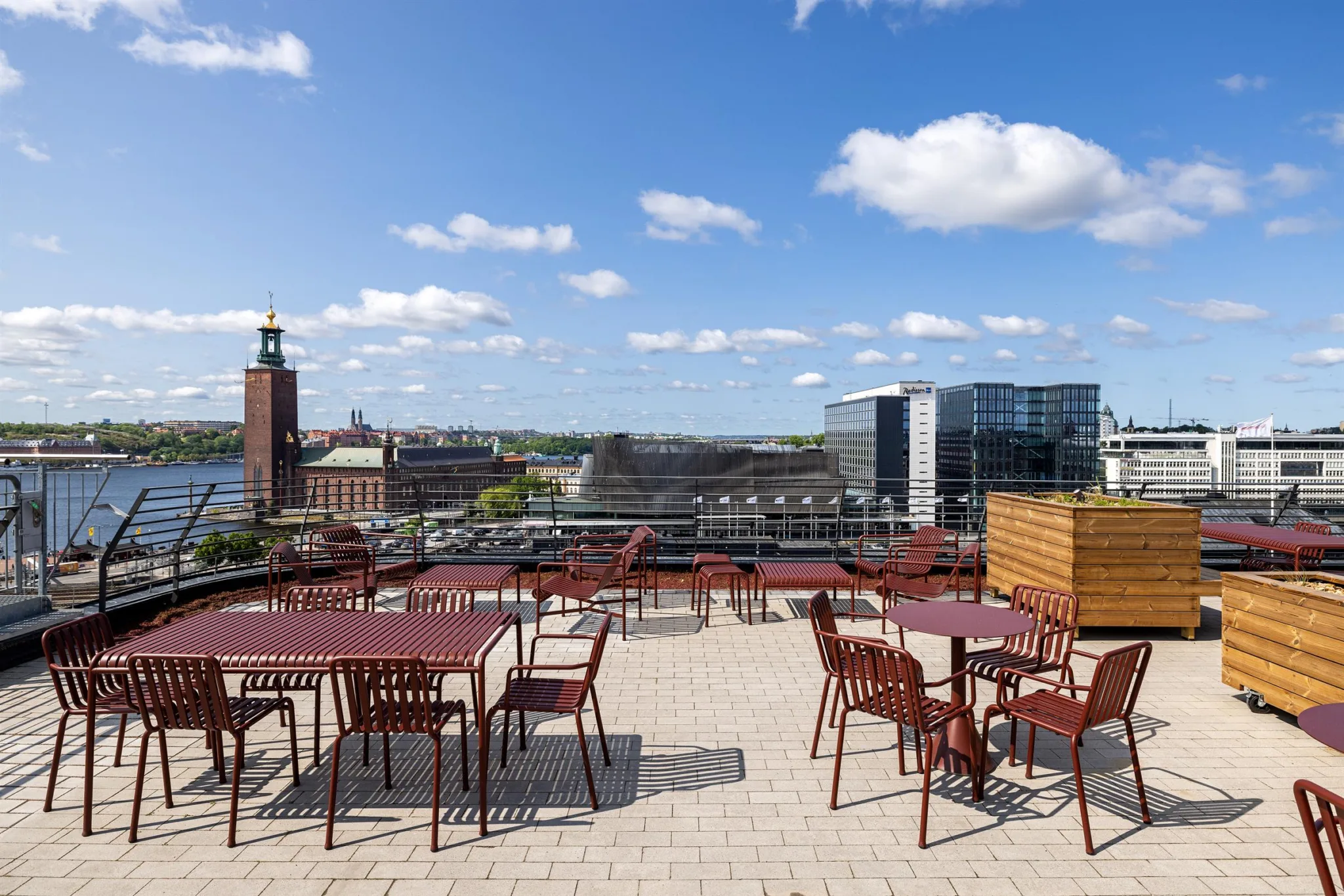 En takterrass med röda bord och stolar, med vy över Stadshuset och moderna byggnader.
