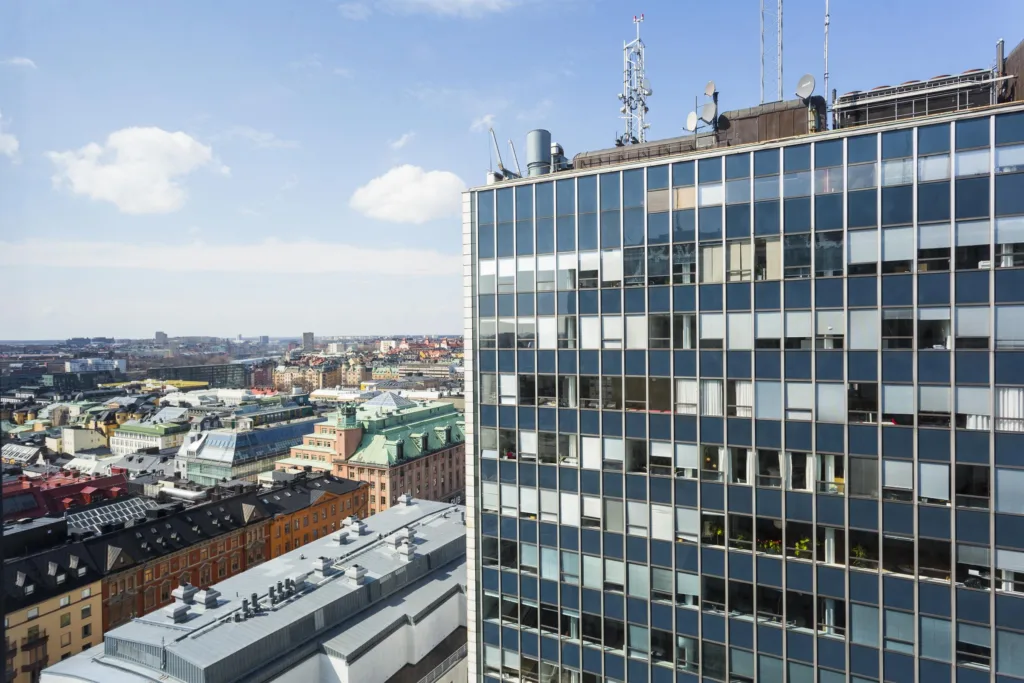 Höghus med glasfasad, antenner på taket och stadsutsikt i bakgrunden.