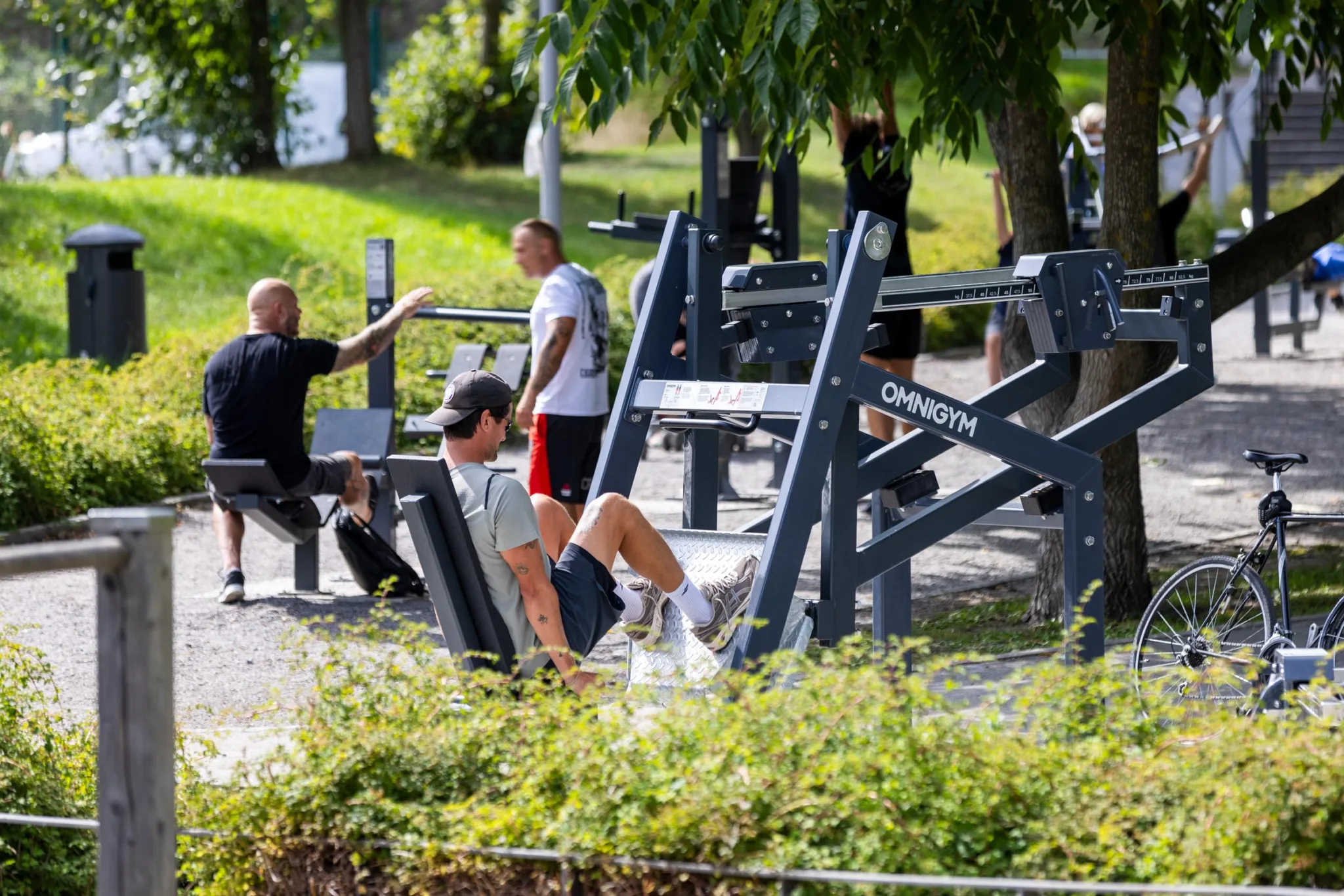 Utomhusgym med flera personer som tränar på olika träningsredskap, omgivna av grönska.
