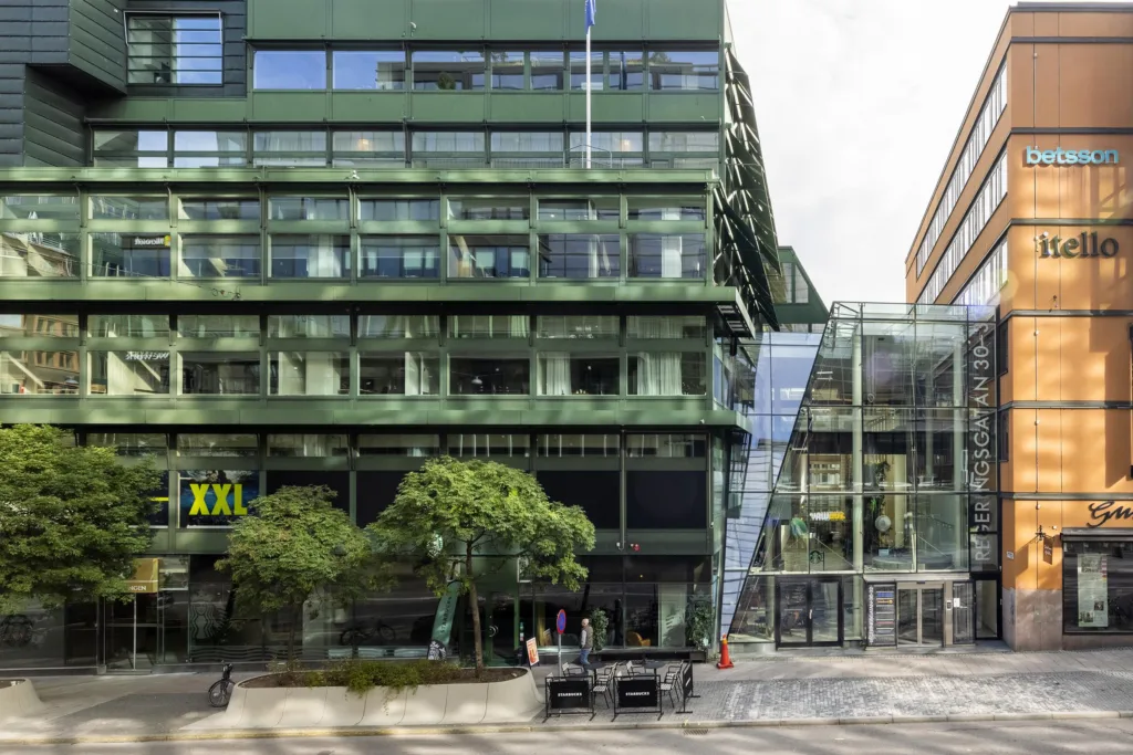 En byggnad med gröna glasfasader, en bred trappa och en XXL-skylt, omgiven av träd och trottoar.