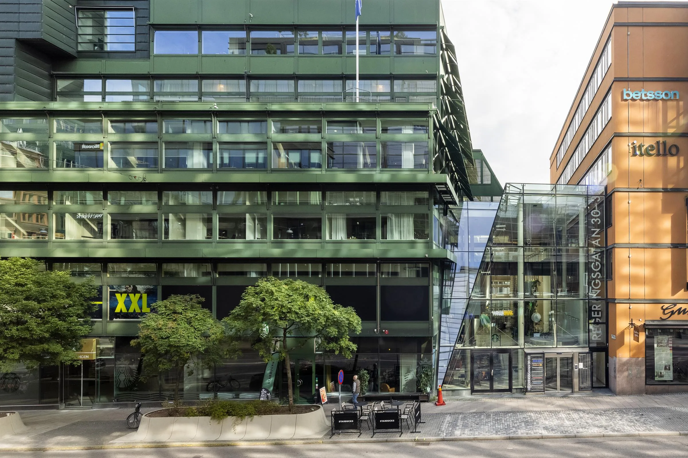 En byggnad med gröna glasfasader, en bred trappa och en XXL-skylt, omgiven av träd och trottoar.