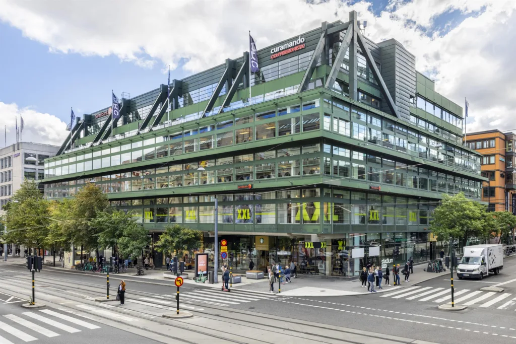 En modern byggnad med glasfasad och gröna detaljer, omgiven av stadsgator och gående människor.