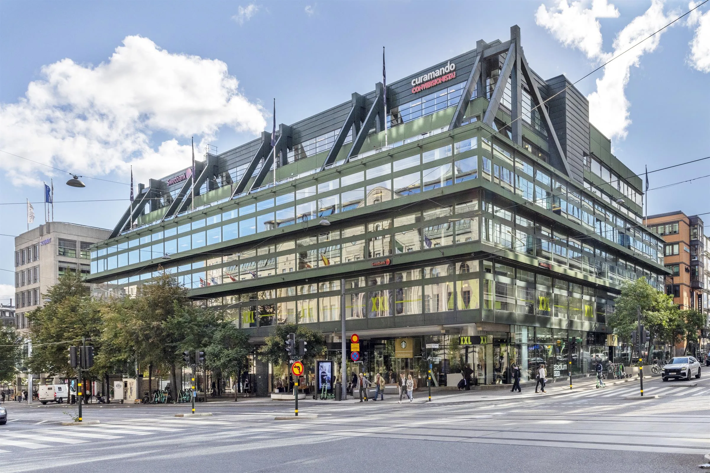 En stor byggnad med glasfasader och gröna takdetaljer, omgiven av träd och stadstrafik.