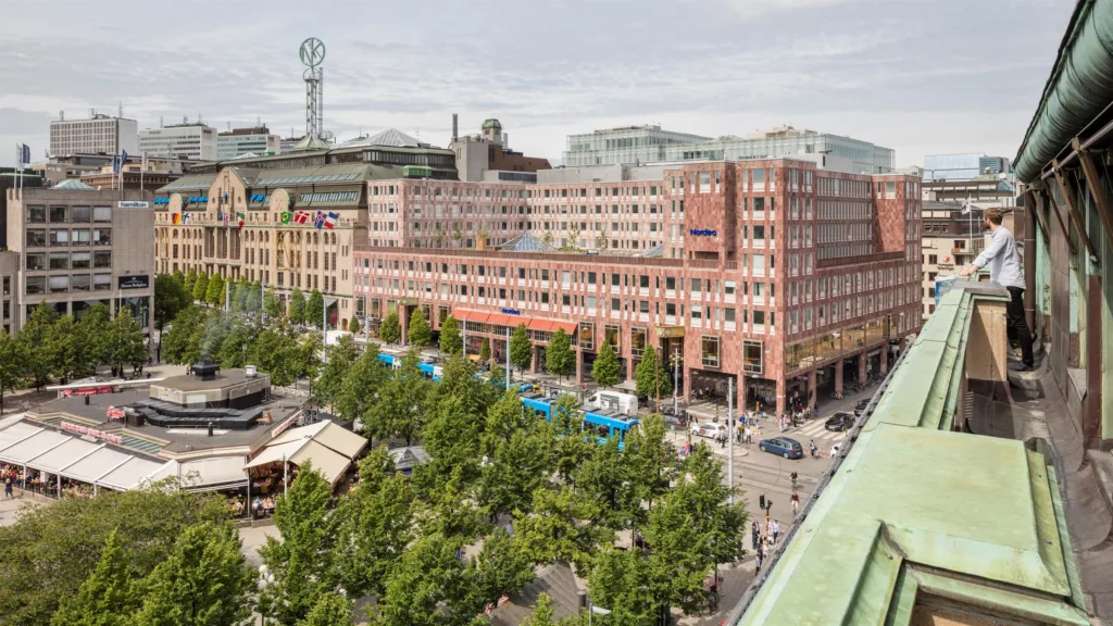 Ett stadsområde med träd, marknadsstånd och en stor byggnad i tegel med flera våningar och skyltar.