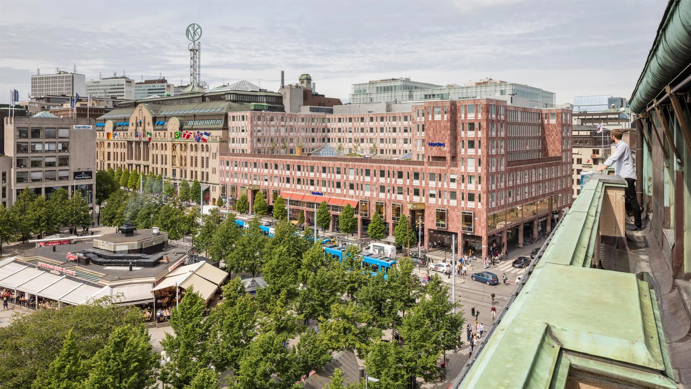 Ett stadsområde med träd, marknadsstånd och en stor byggnad i tegel med flera våningar och skyltar.
