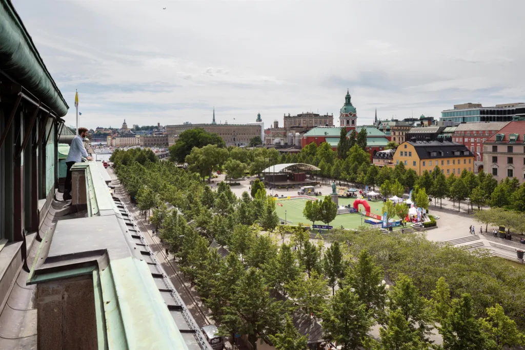 En utsikt över en grön park med träd, färgglada tält och byggnader i bakgrunden.