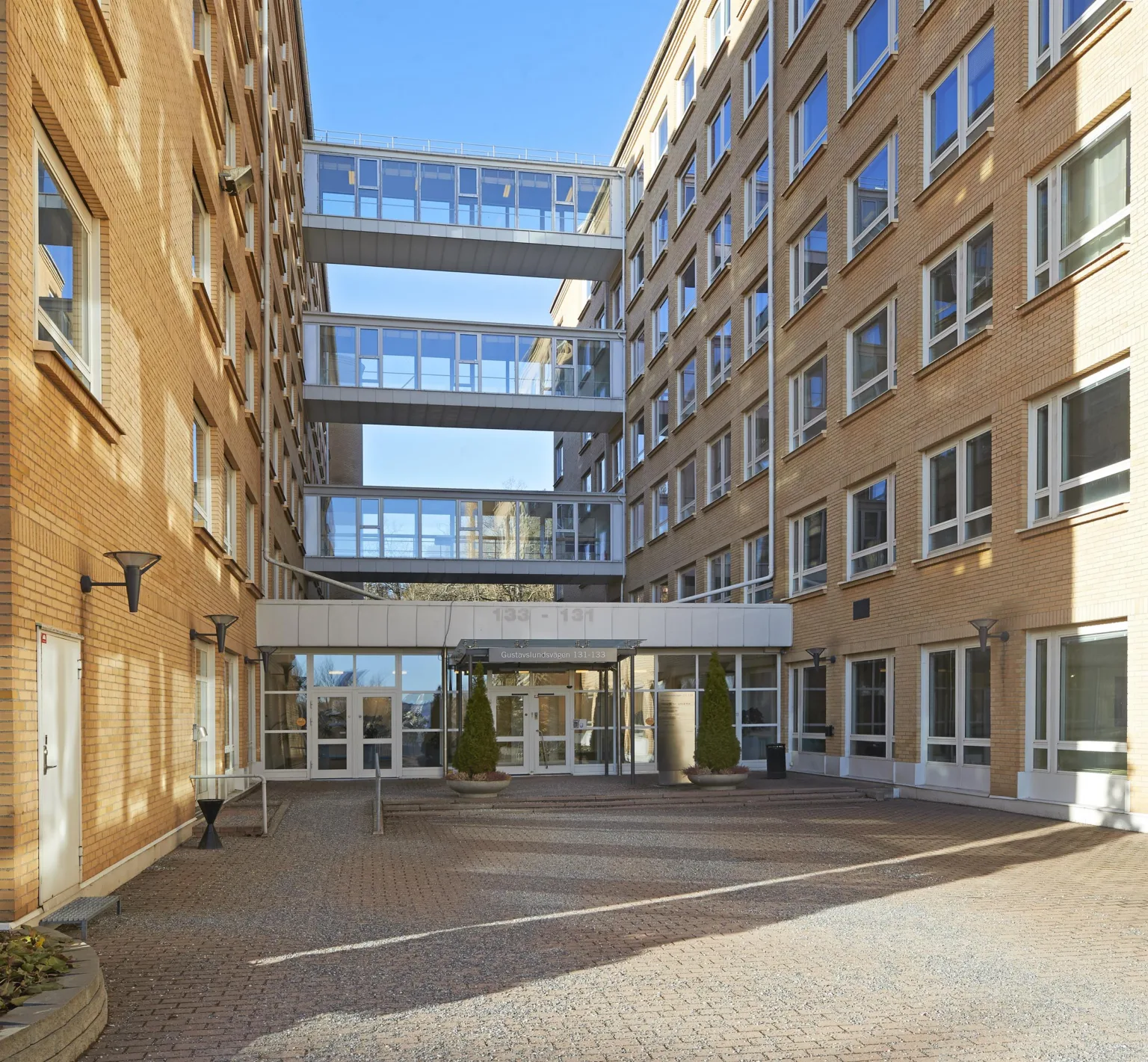 En byggnad med gul fasad och glasgångar som förbinder två flyglar, med en stenlagd innergård och klarblå himmel i bakgrunden.