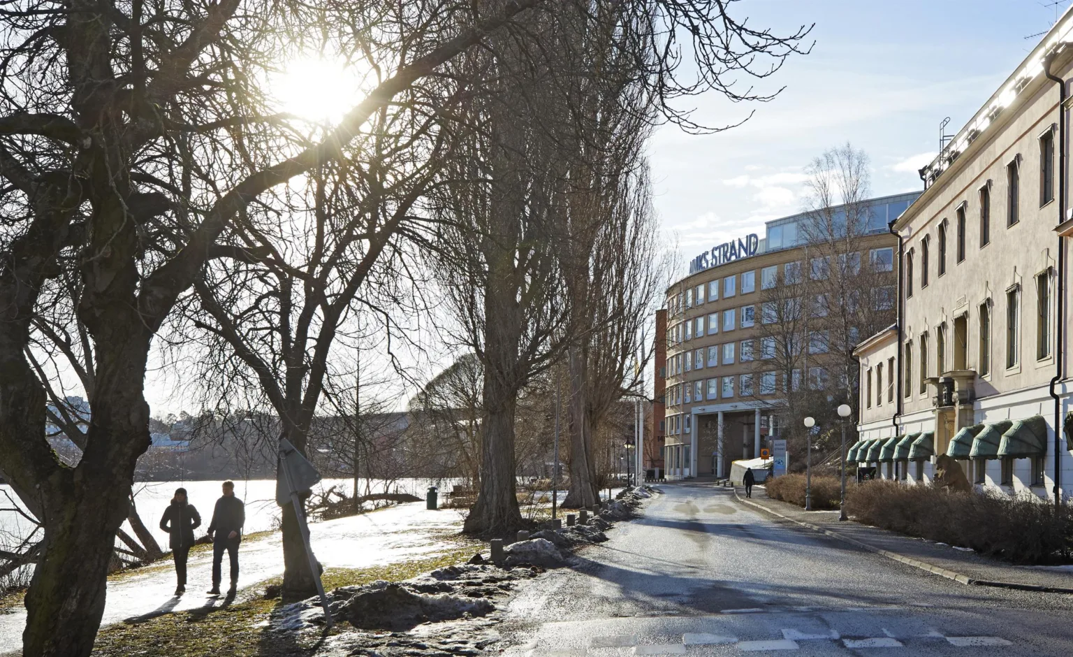 En gångväg med snöfläckar och träd längs sidorna, där solen skiner genom grenarna, med en byggnad i bakgrunden.