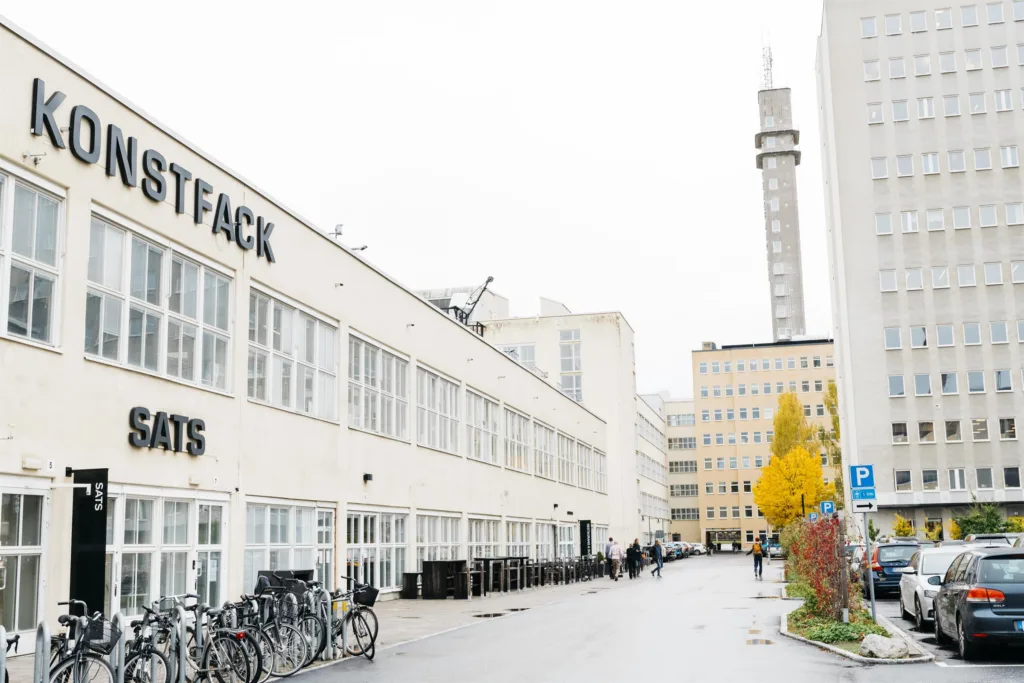 En vit byggnad med texten "STOCKHOLMS KONSTFACK" på fasaden, cyklar parkerade längs vägen och en tornliknande byggnad i bakgrunden.