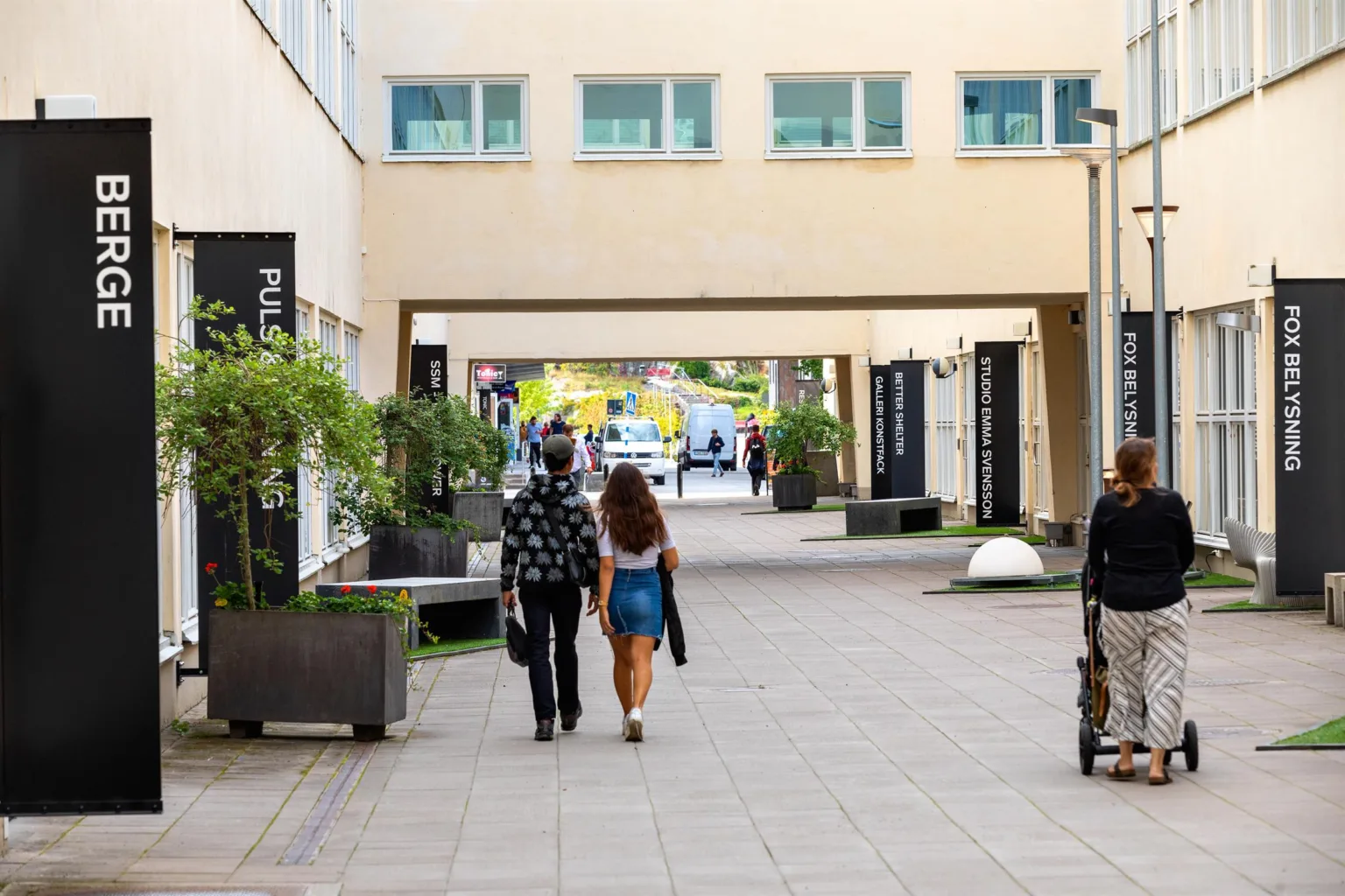 En gångväg mellan byggnader där människor promenerar, med gröna växter i krukor längs sidorna.
