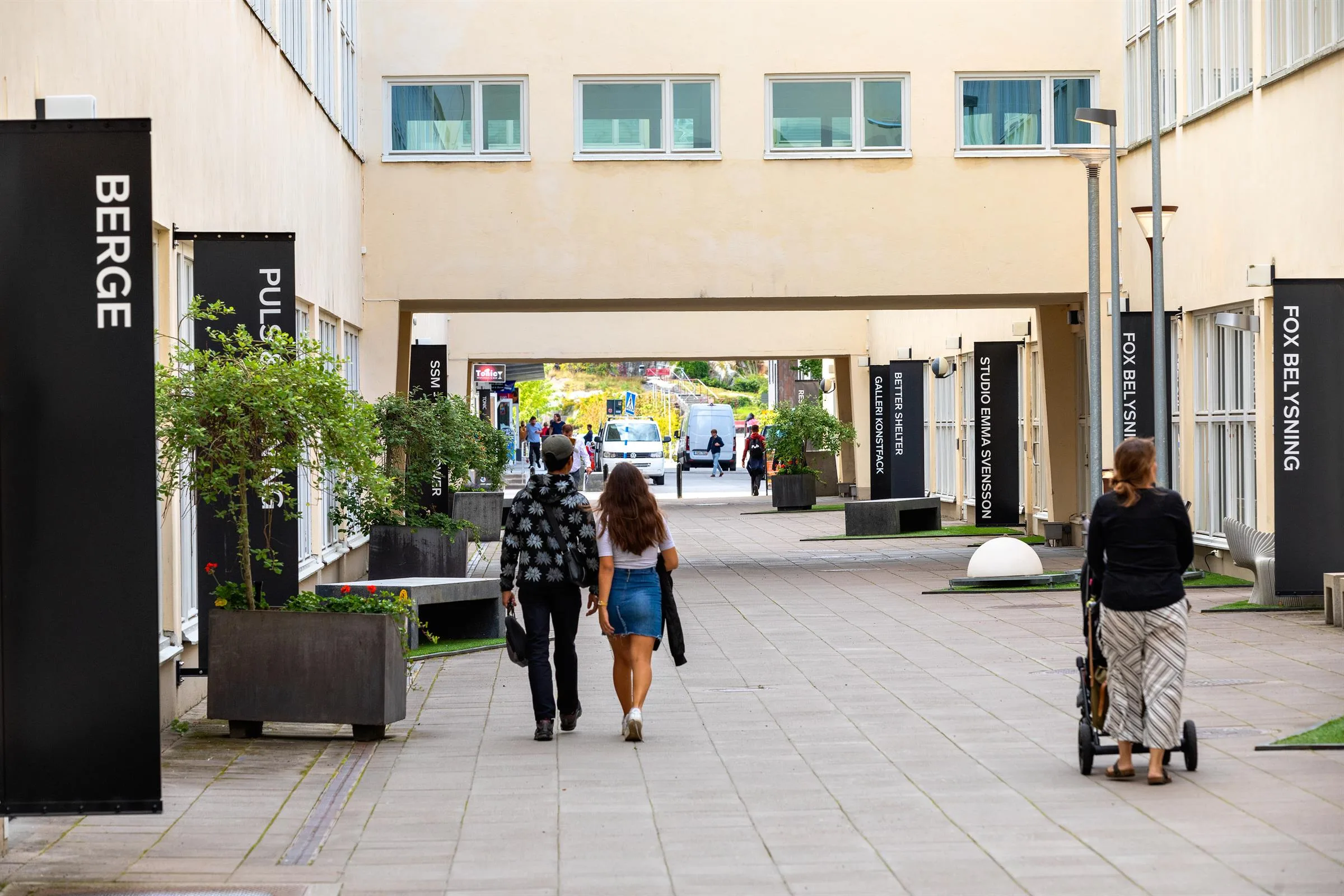 En gångväg mellan byggnader där människor promenerar, med gröna växter i krukor längs sidorna.