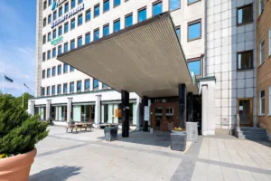 Entrén till en modern byggnad med en bred takskärm, flera fönster och blomkrukor placerade utanför.