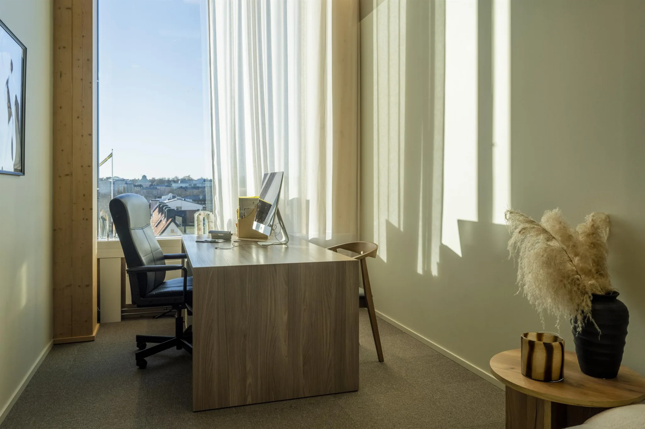 Ett kontor med ett stort skrivbord, en skrivbordsstol och stora fönster med utsikt över en stad.