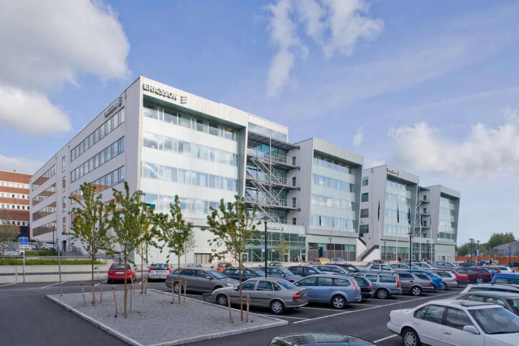En vit kontorsbyggnad med glasfönster, parkeringsplats med flera bilar i förgrunden.