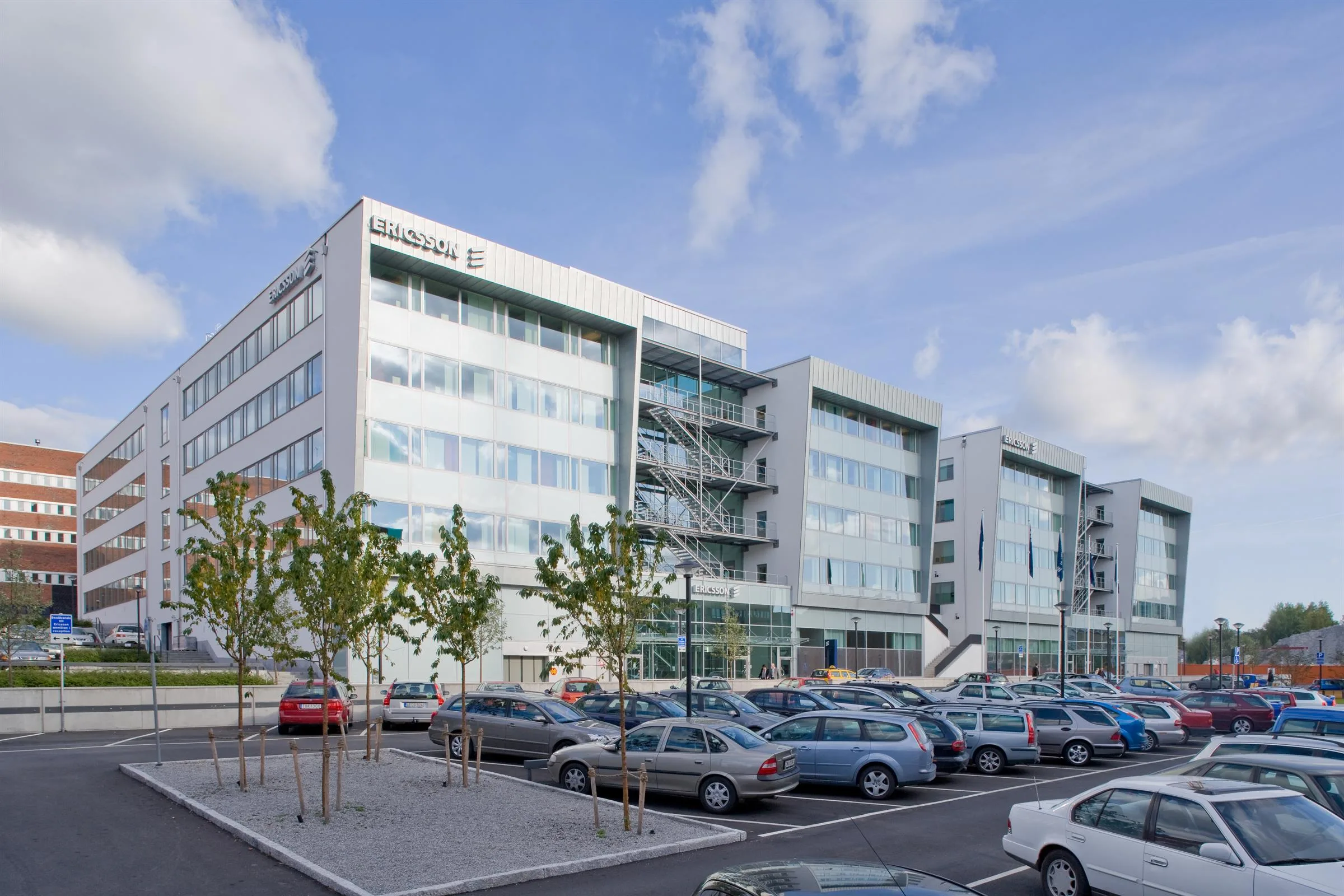 En vit kontorsbyggnad med glasfönster, parkeringsplats med flera bilar i förgrunden.