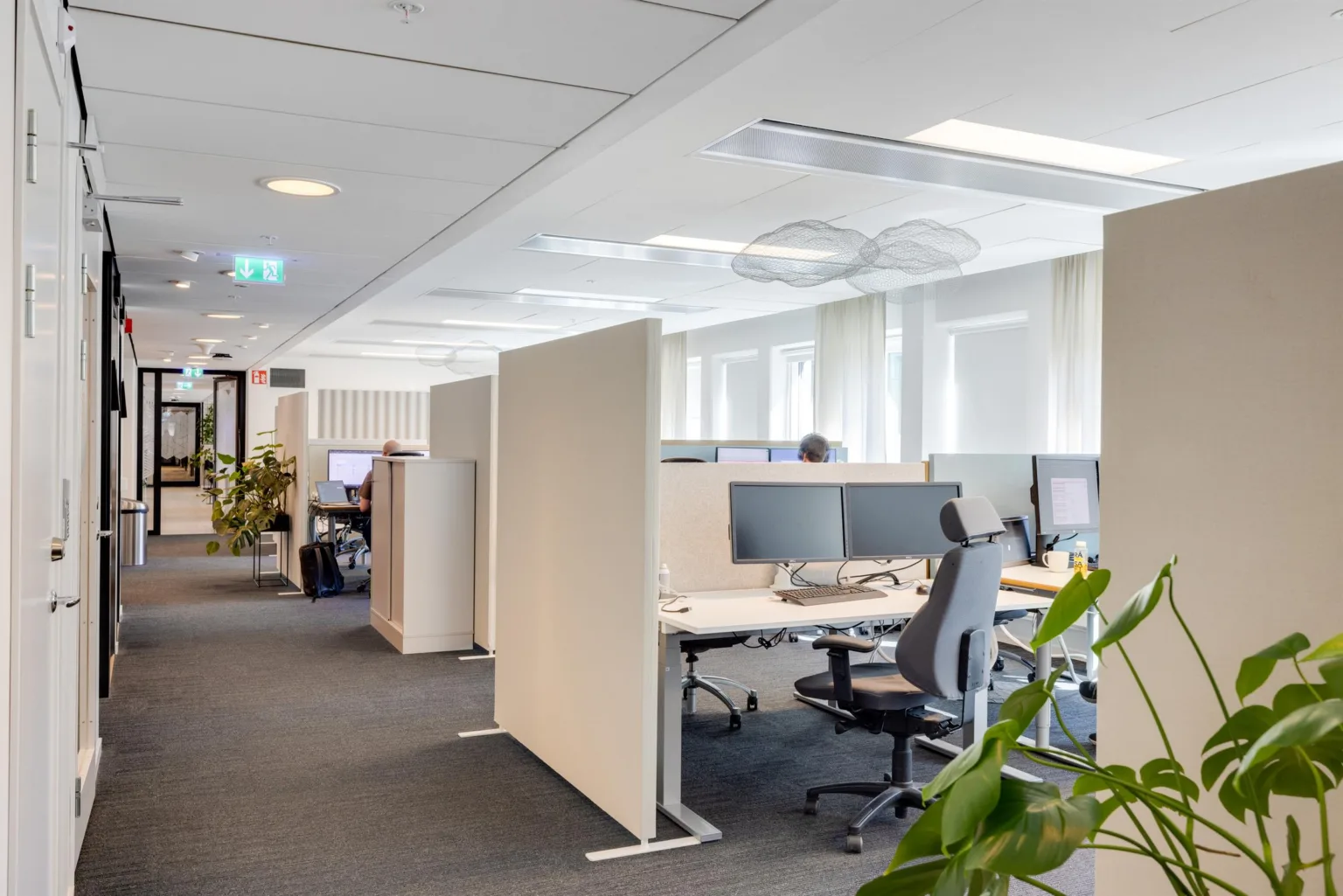 En öppen kontorsmiljö med arbetsstationer, skrivbord och stolar, omgiven av växter och naturligt ljus.