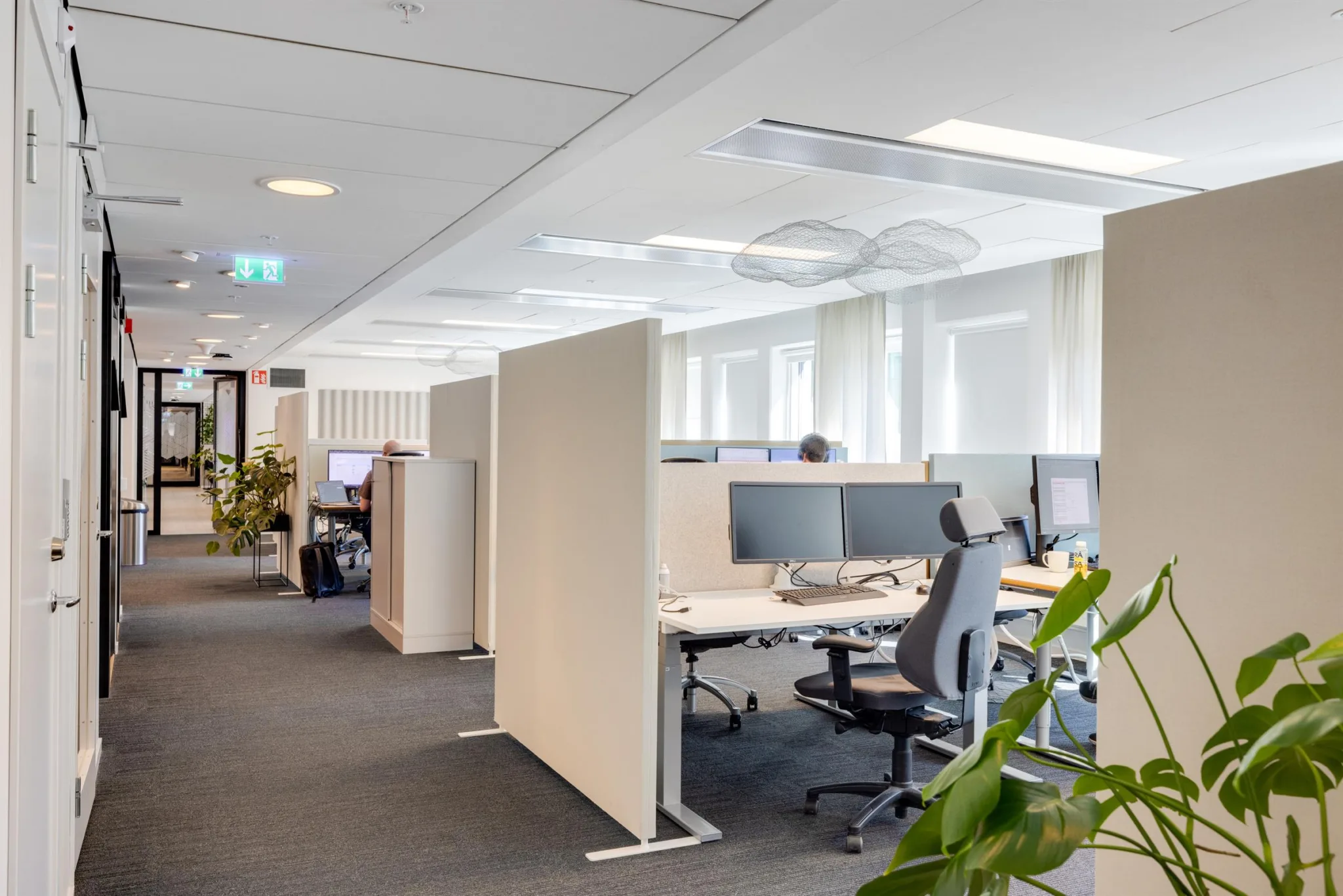 En öppen kontorsmiljö med arbetsstationer, skrivbord och stolar, omgiven av växter och naturligt ljus.