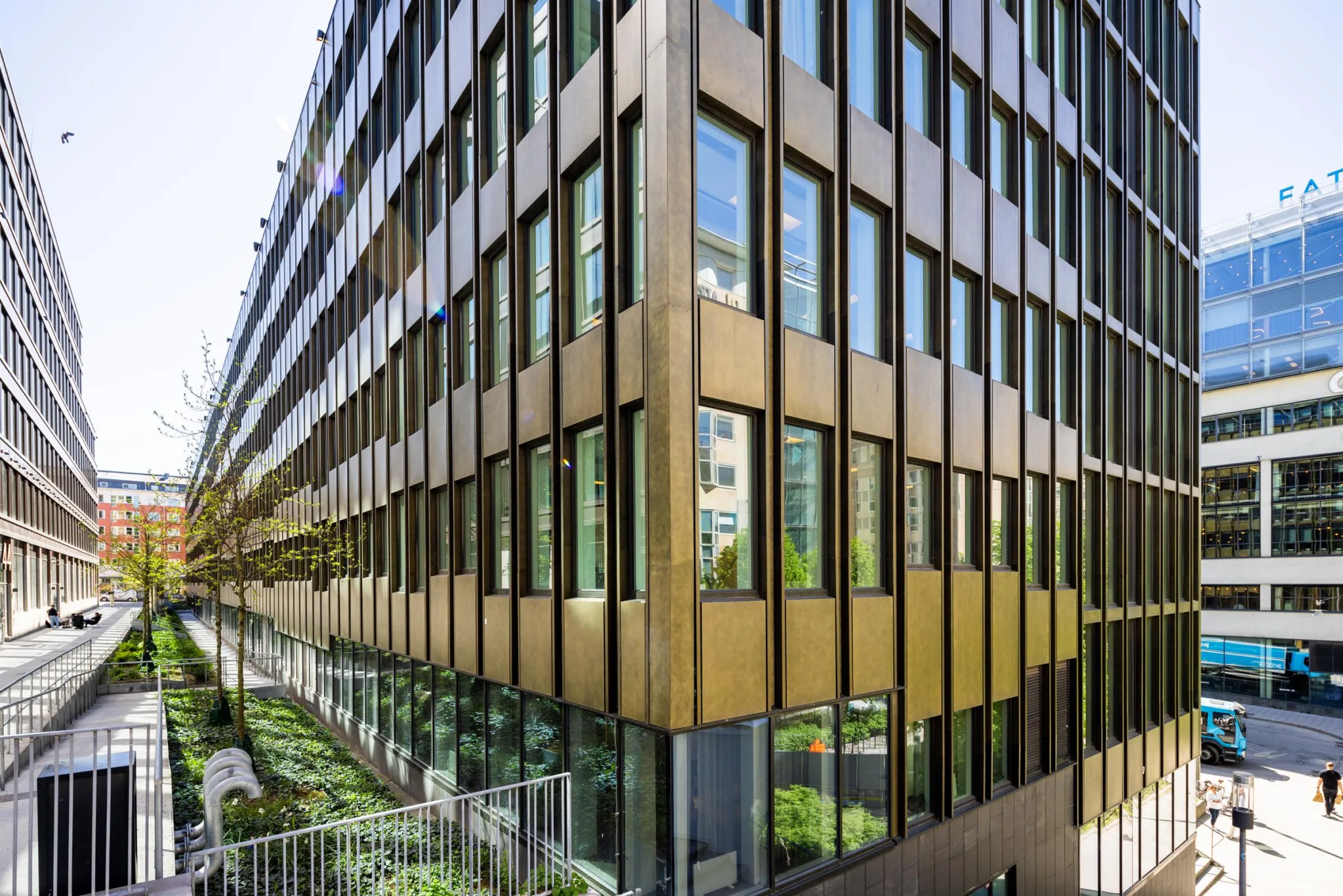 En modern byggnad med glasfönster och metallisk fasad, omgiven av grönområden och gångstigar.