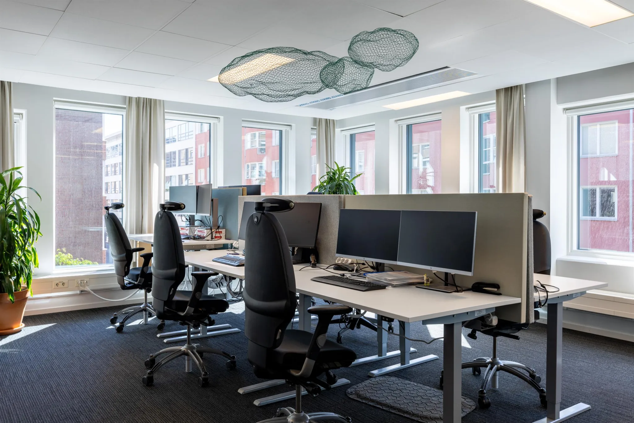 En kontorsmiljö med flera skrivbord, kontorsstolar och datorskärmar, med stora fönster som ger mycket naturligt ljus.