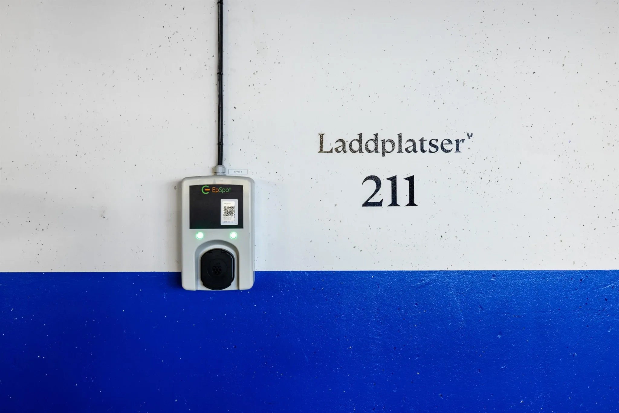 En laddstation för elfordon med texten "Laddplatser 211" på en vägg målad i vitt och blått.