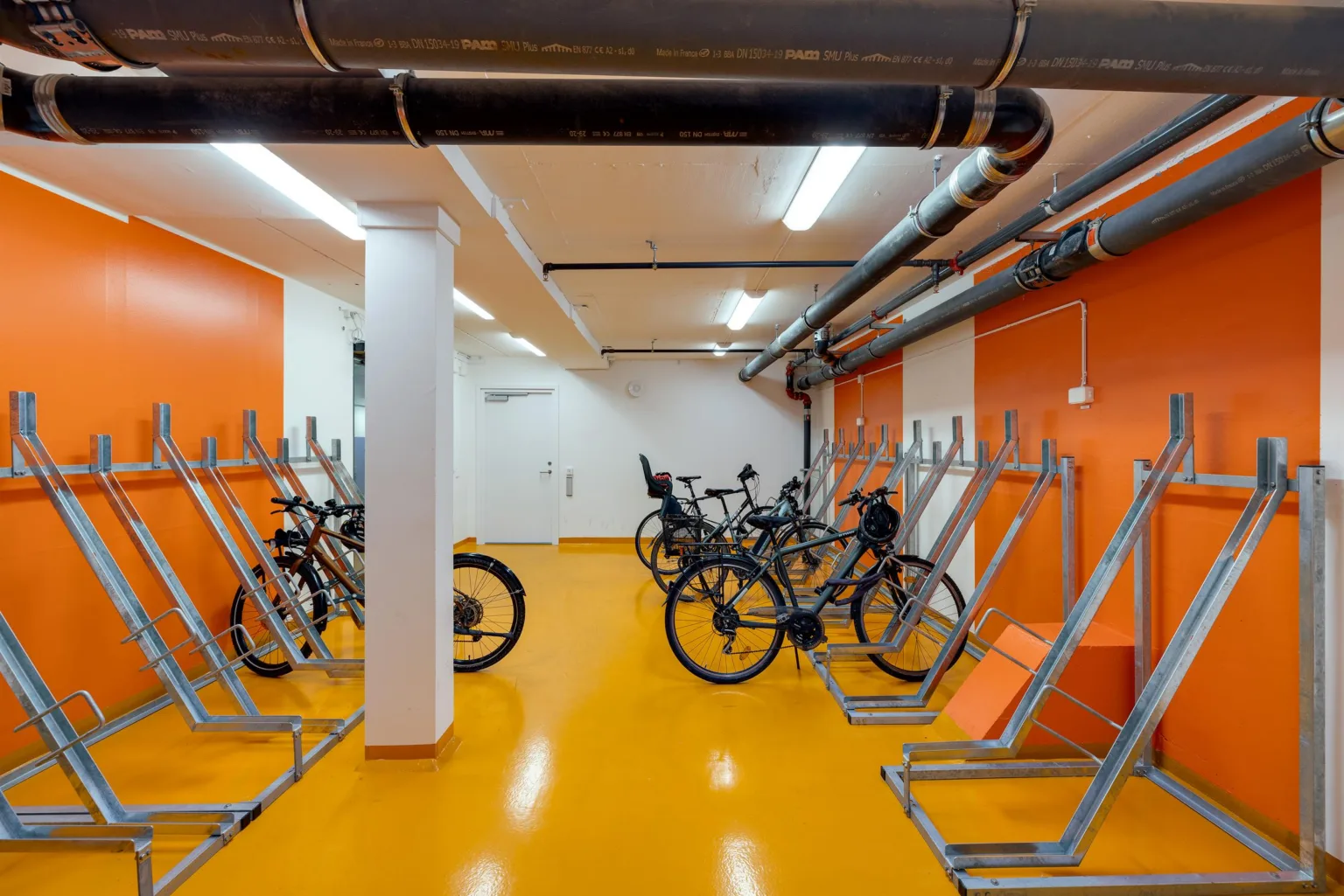 En cykelparkering inomhus med orange golv, cykelställ och flera parkerade cyklar.
