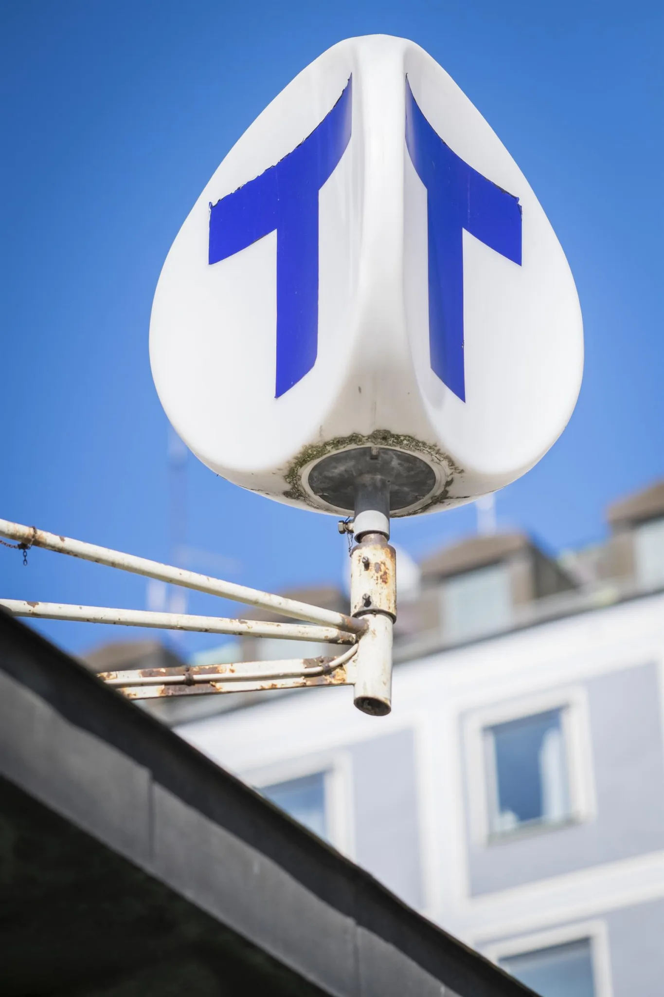 En vit vägskylt med två blå pilar som pekar uppåt, monterad på en stolpe med en byggnad och klarblå himmel i bakgrunden.
