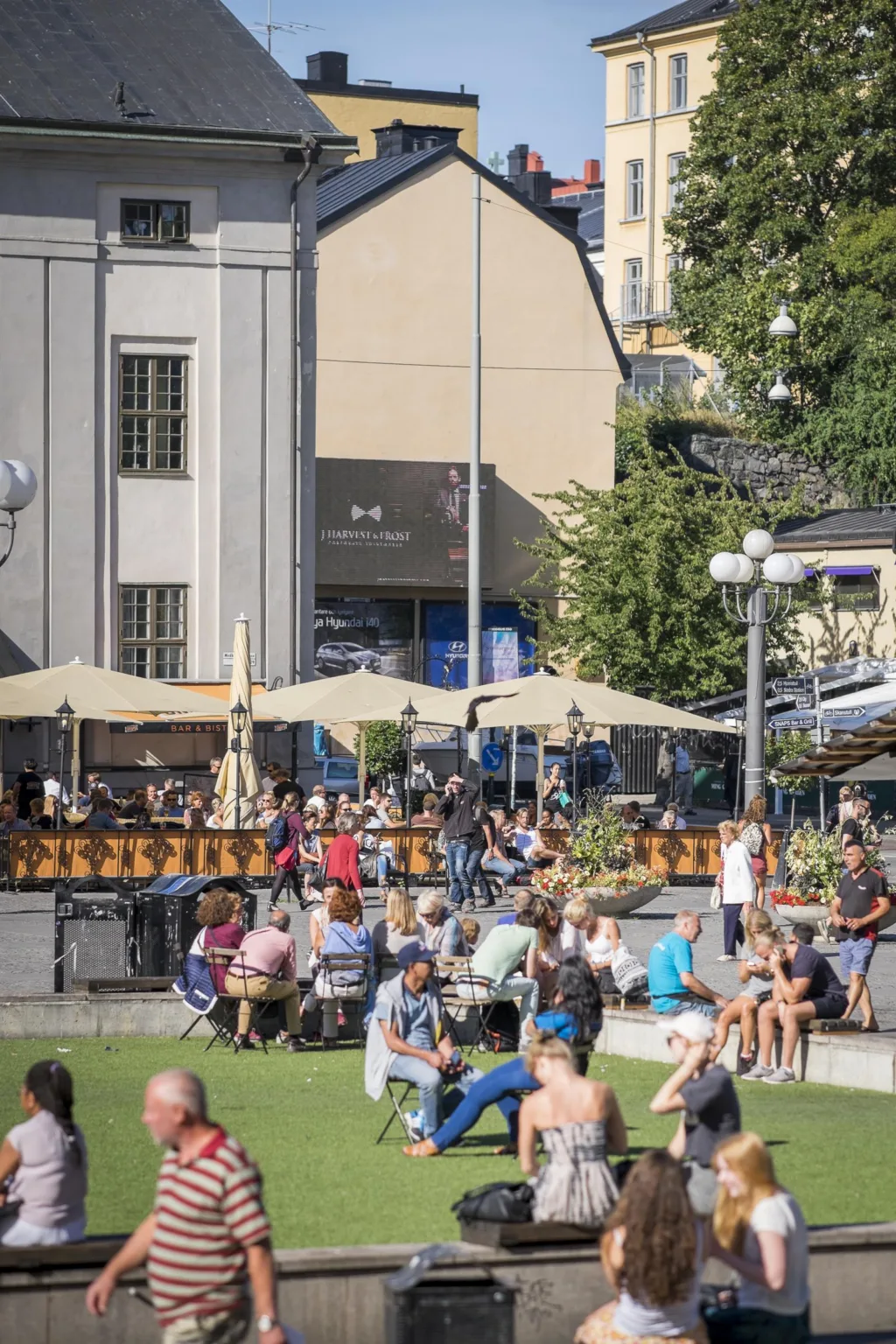 En grupp människor sitter och står vid ett torg med cafébord och parasoller, omgivna av byggnader och grönska.