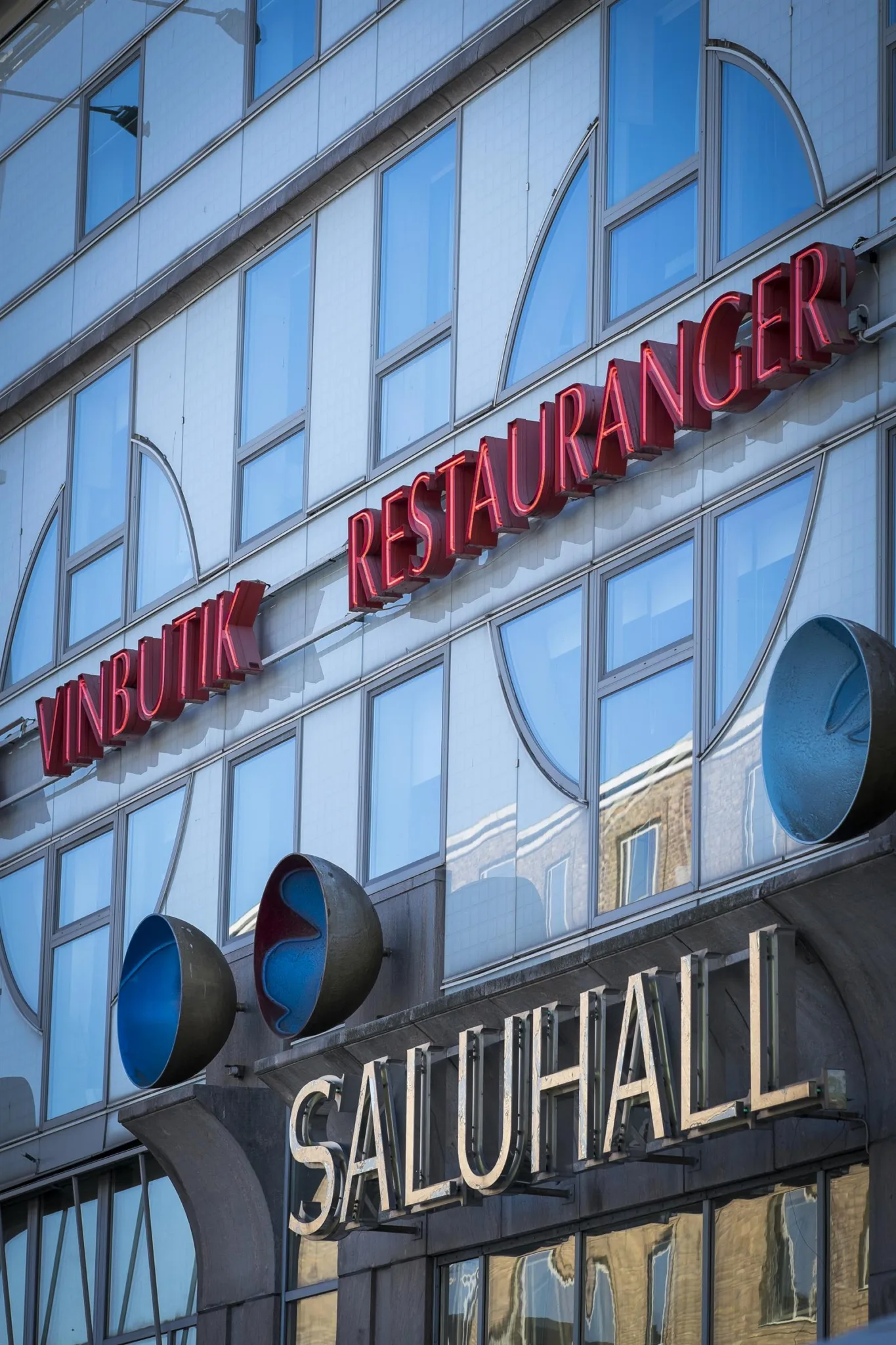 Fasaden på en byggnad med texten "Vinbutik Restauranger Saluhall" i röda och vita bokstäver.