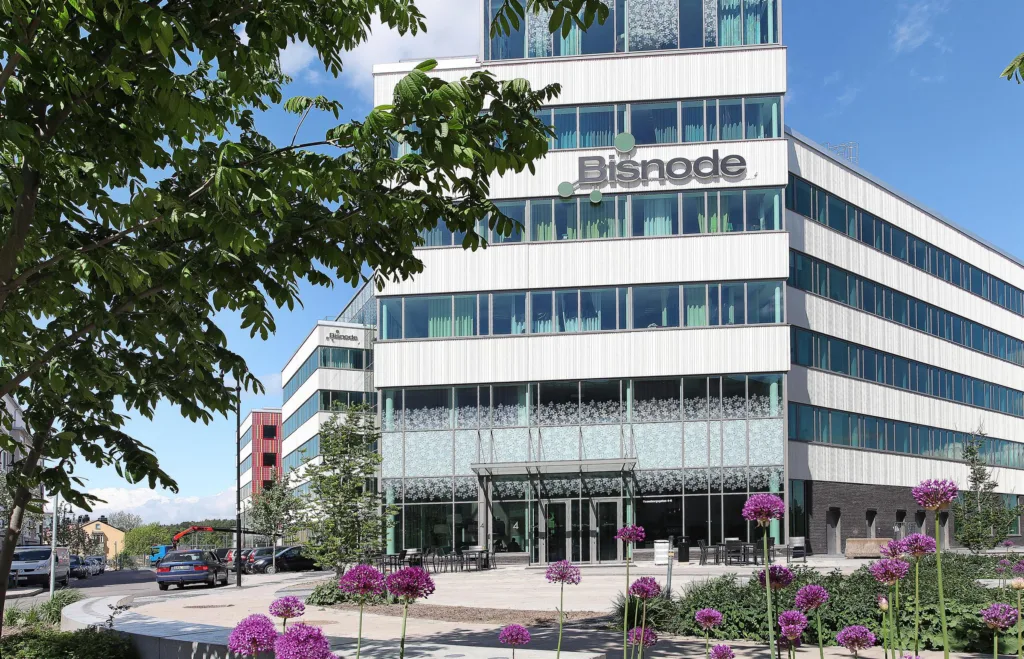 Glasbyggnad med texten "Bisnode", omgiven av blommor och träd i en grönskande omgivning.
