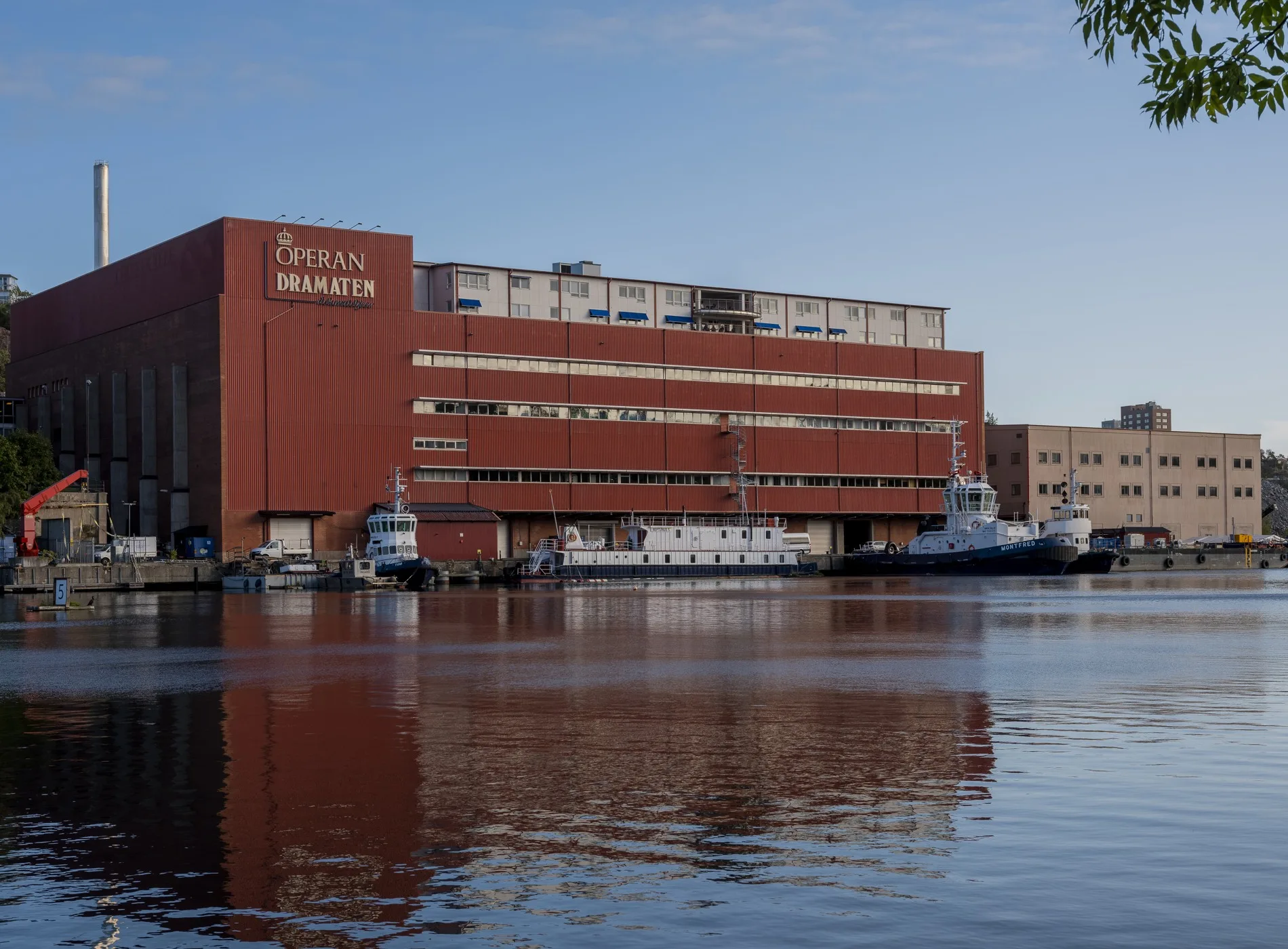 En stor röd byggnad med texten "Campus Haninge" vid en vattenyta, med båtar och reflektioner i vattnet.
