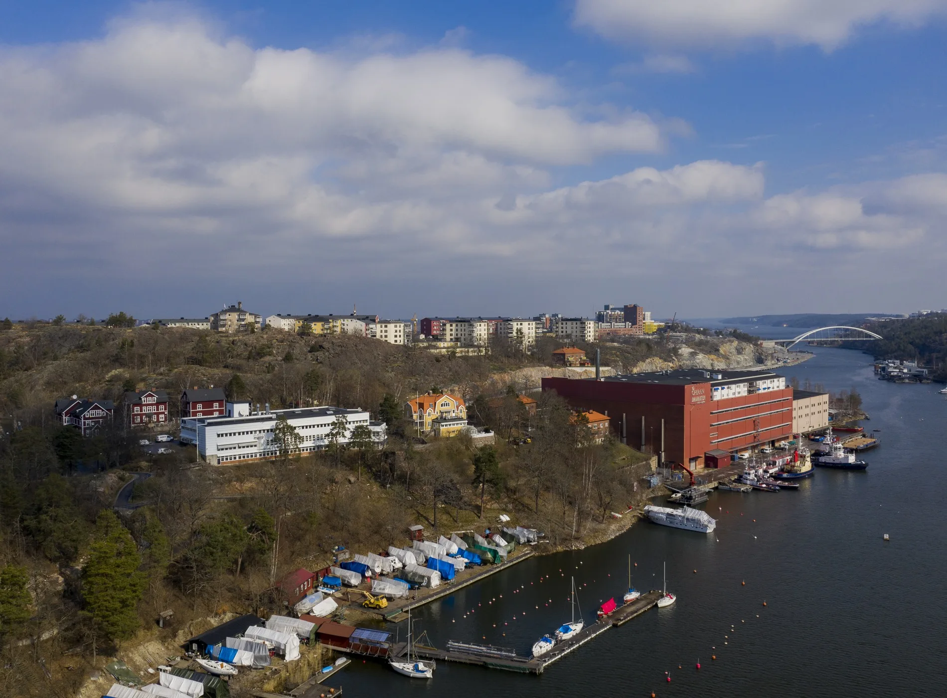 En flygbild över en havsvik med båtar och en röd byggnad, samt hus och grönska i bakgrunden.