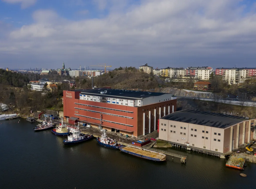 En röd byggnad vid en havsvik med båtar och omkringliggande stadsbebyggelse.